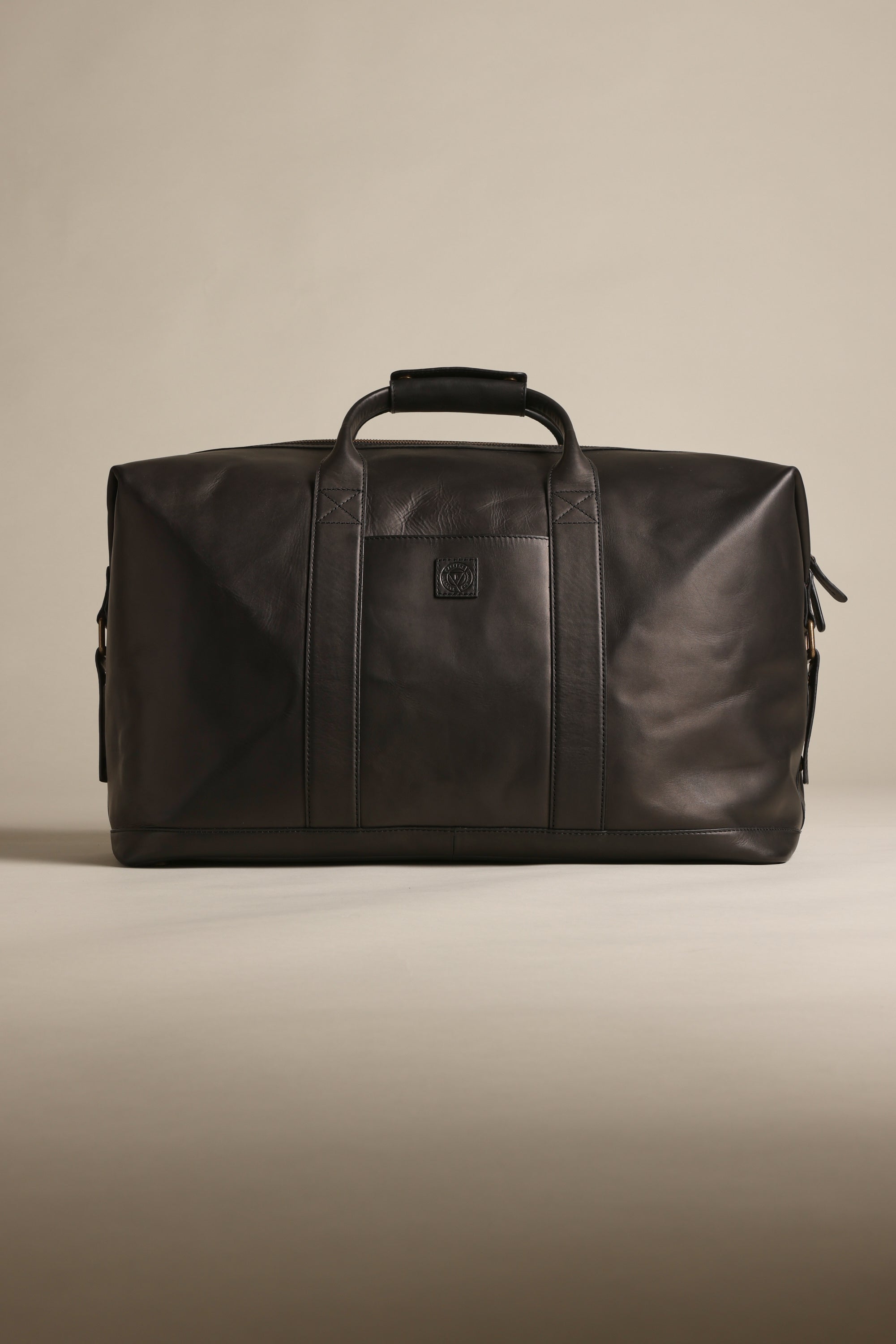 The Barkers Costello Leather Weekender, a black premium leather bag with two handles and a shoulder strap, features a minimalist design and small embossed logo on the front. Displayed against a neutral beige background.