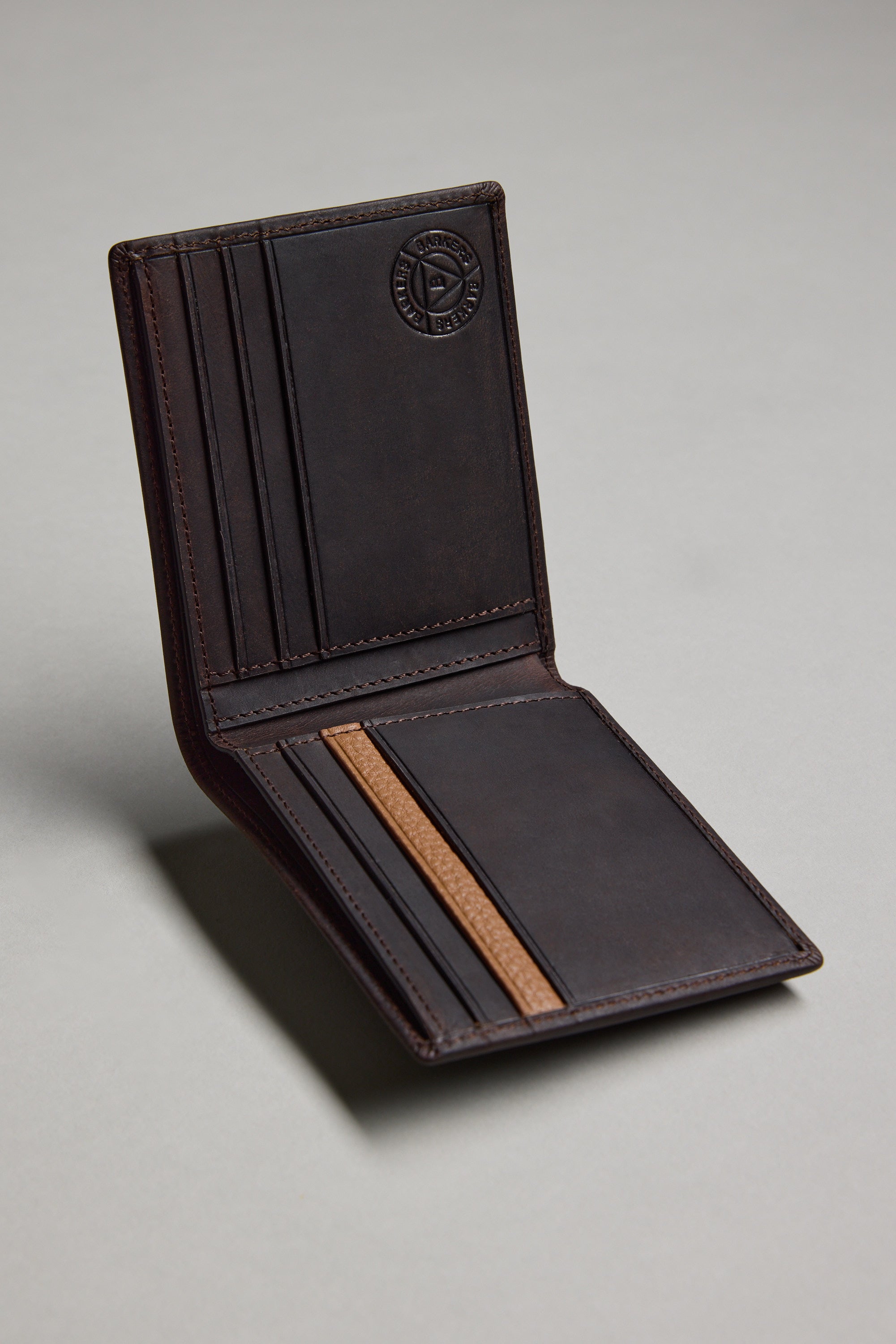 The Barkers Harris Leather Fold Wallet is open, showing multiple card slots and a tan accent strip inside. An embossed circular logo appears on the inner flap. The slim wallet's dark brown leather stands out against a plain light gray background.