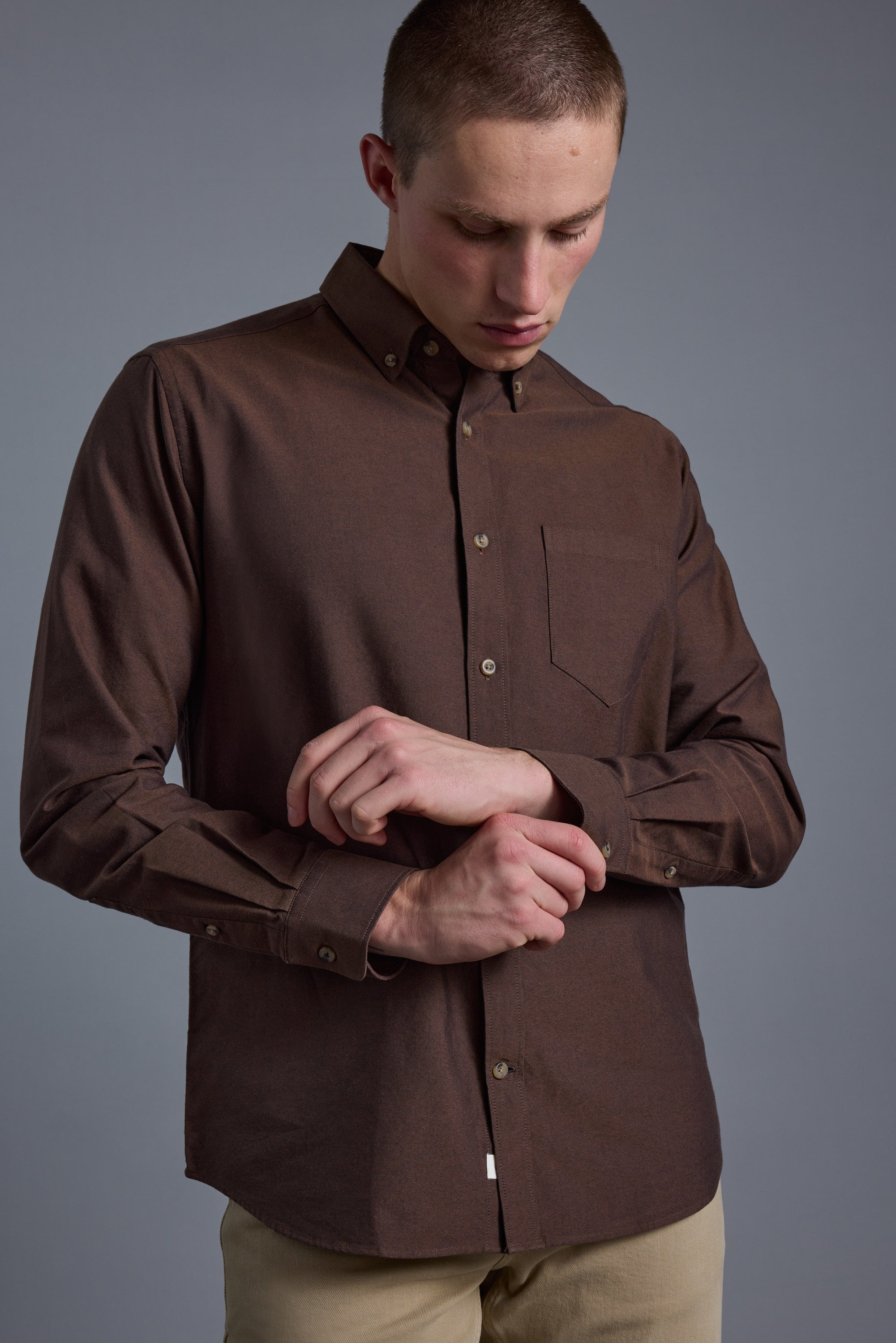 Wearing the Barkers Thatcher Oxford Shirt with a chest pocket and beige pants, a person adjusts their cuff while looking down, posed against a plain gray background.