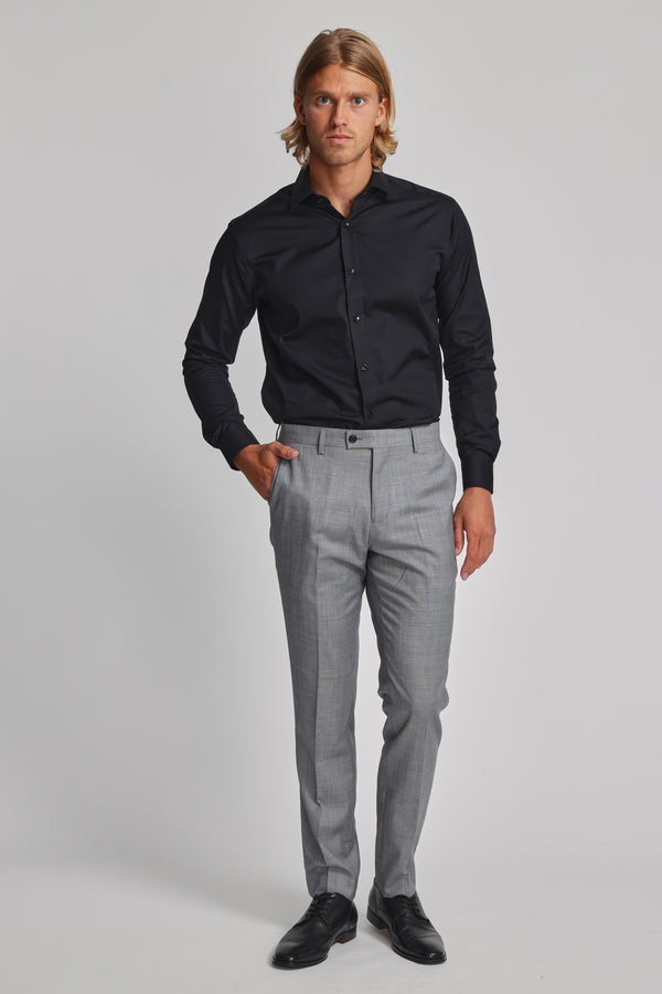 A man stands against a plain background wearing the Barkers Tribeca Reg Cuff Shirt. He pairs the tailored, wrinkle-free black dress shirt with light gray dress pants and black dress shoes, one hand in his pocket and a neutral expression on his face.