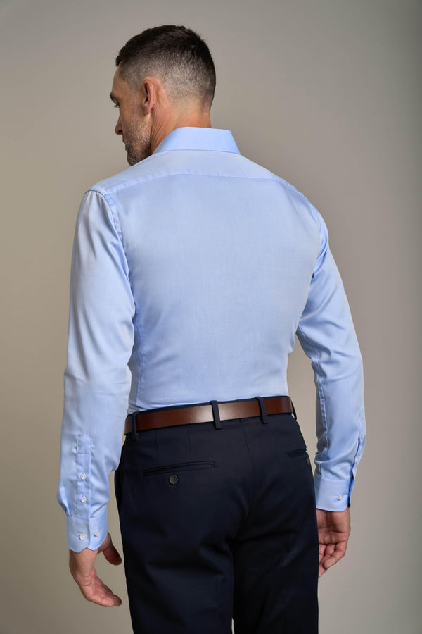 A man with short hair is shown from the back, wearing the Barkers Tribeca Reg Cuff Shirt in light blue, tucked into black dress pants with a brown belt, standing against a plain beige background.