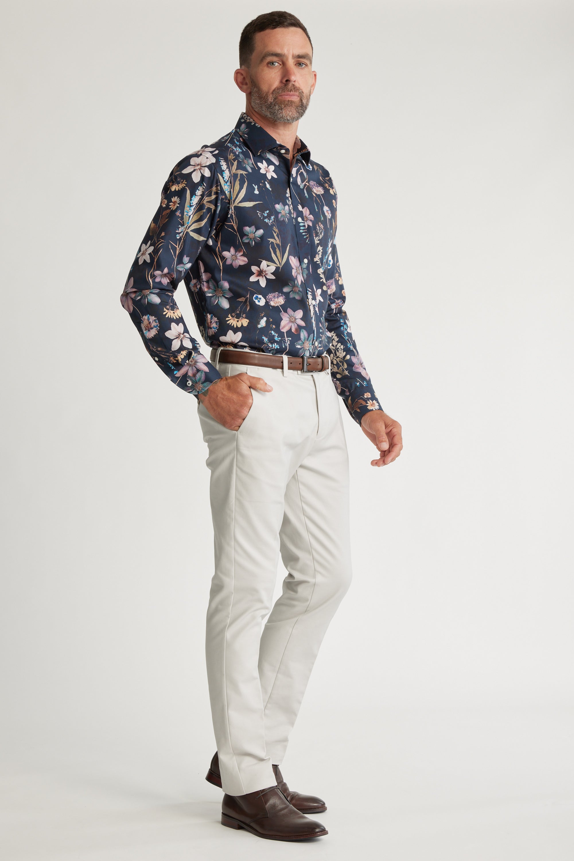 A man with short hair and a beard stands confidently against a white background wearing the Barkers Apothecary AOP Business Shirt in dark blue floral, paired with light grey pants, a brown belt, and brown dress shoes.