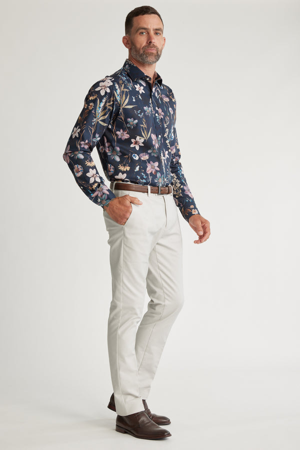 A man with short hair and a beard stands confidently against a white background wearing the Barkers Apothecary AOP Business Shirt in dark blue floral, paired with light grey pants, a brown belt, and brown dress shoes.