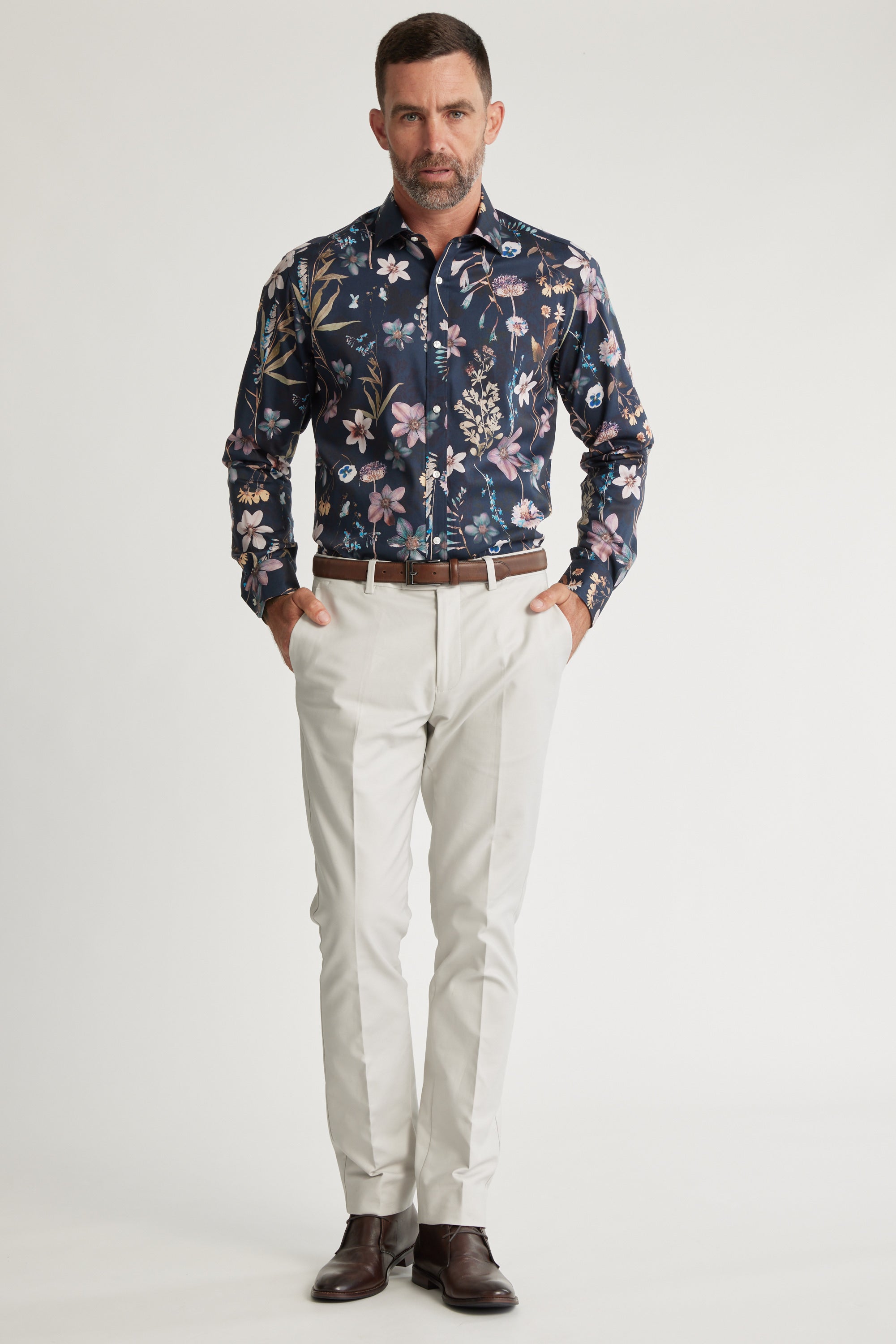 A man with short dark hair and a beard stands against a white background wearing a Barkers Apothecary AOP Business Shirt, light trousers, a brown belt, and brown dress shoes. His hands are in his pockets.