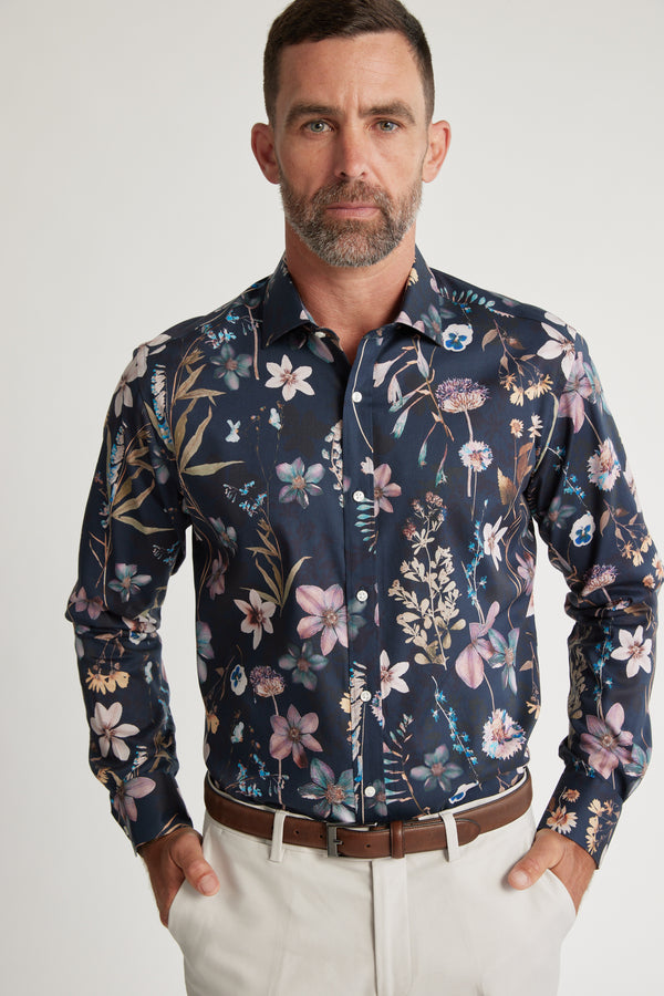 A man with short hair and a beard wears a dark, long-sleeve Barkers Apothecary AOP Business Shirt and beige pants. He stands with his hands in his pockets against a plain, light background.