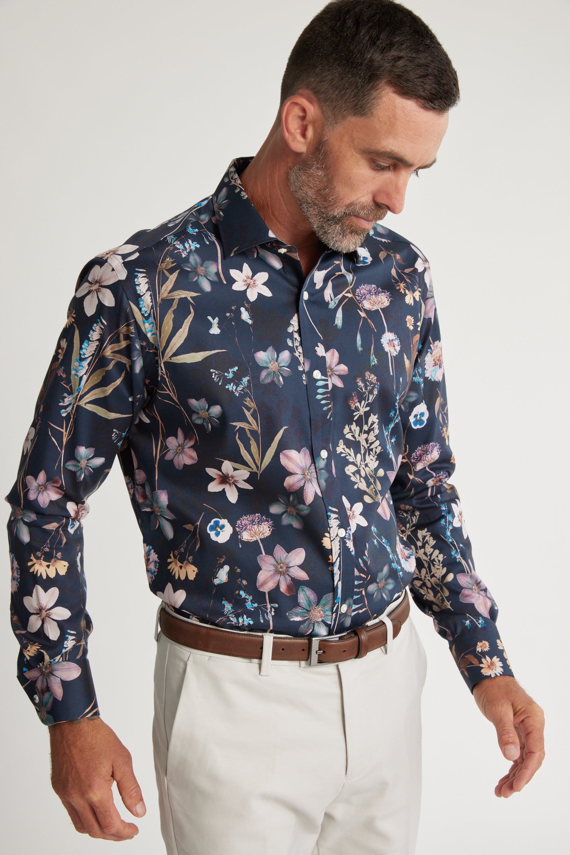 A man wearing a dark blue Barkers Apothecary AOP Business Shirt with a colorful floral pattern, paired with light pants and a brown belt. This classic fit shirt is crafted from responsibly sourced cotton.