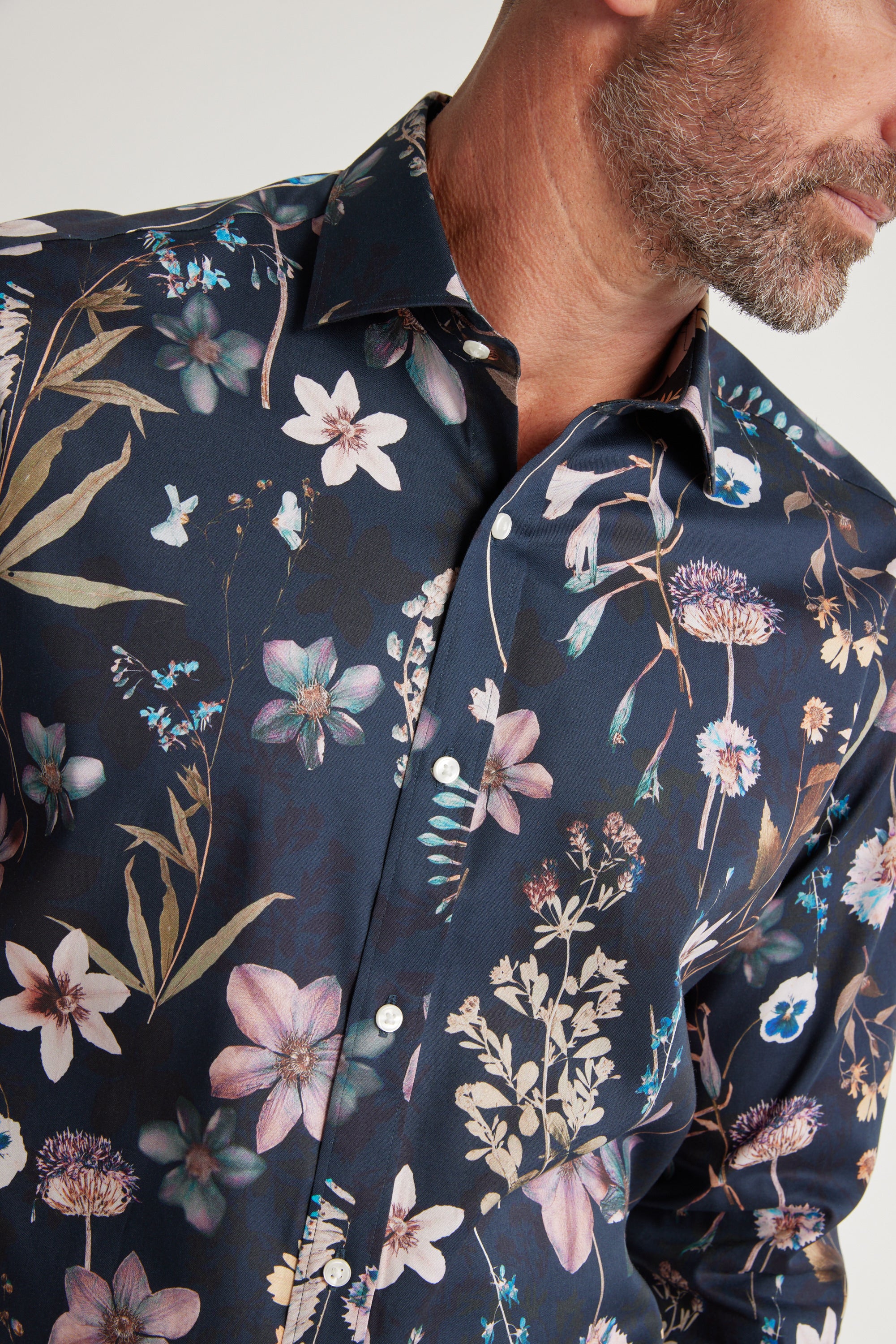 A man with a short beard wears the Barkers Apothecary AOP Business Shirt, a classic-fit, responsibly sourced cotton shirt in dark blue with a vivid floral print in pink, purple, beige, and green. Cropped at the shoulders.
