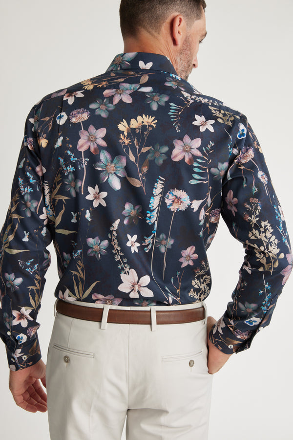 A man with short hair and a trimmed beard stands facing away, wearing the Barkers Apothecary AOP Business Shirt with a colorful floral pattern and light beige pants. The shirt is made from responsibly sourced cotton.
