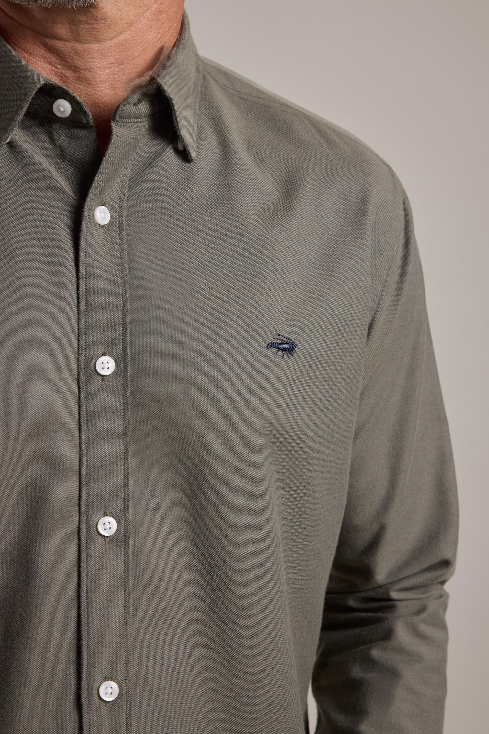 A close-up shows a person wearing the Barkers Maynard Oxford Shirt in olive green, featuring white buttons and a dark blue embroidered logo. Only the lower face, neck, and upper torso are visible.