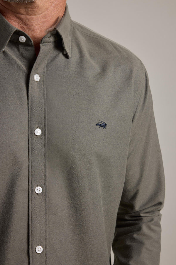 A close-up shows a person wearing the Barkers Maynard Oxford Shirt in olive green, featuring white buttons and a dark blue embroidered logo. Only the lower face, neck, and upper torso are visible.