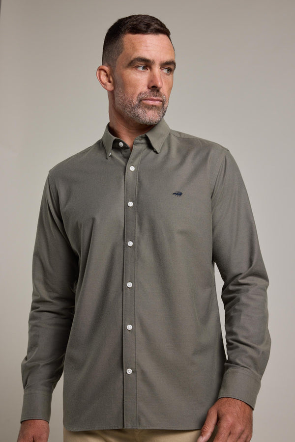 Man with short dark hair and beard wears a buttoned-up, long-sleeve Maynard Oxford Shirt by Barkers in olive green, looking to his left against a plain light background.