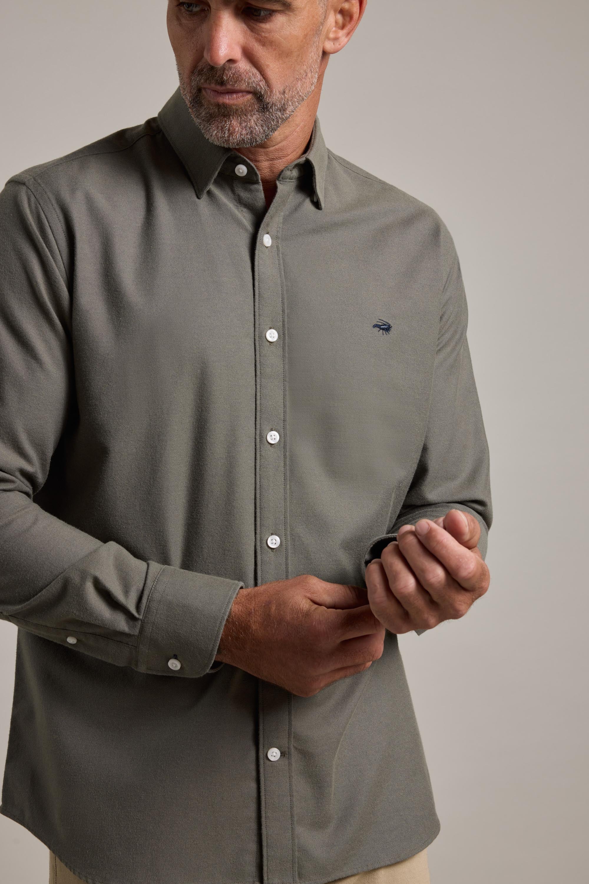 A man with a short beard wears the Barkers Maynard Oxford Shirt, a 100% cotton classic fit in olive green, paired with beige pants. He adjusts his sleeve while facing slightly to the side against a plain background.