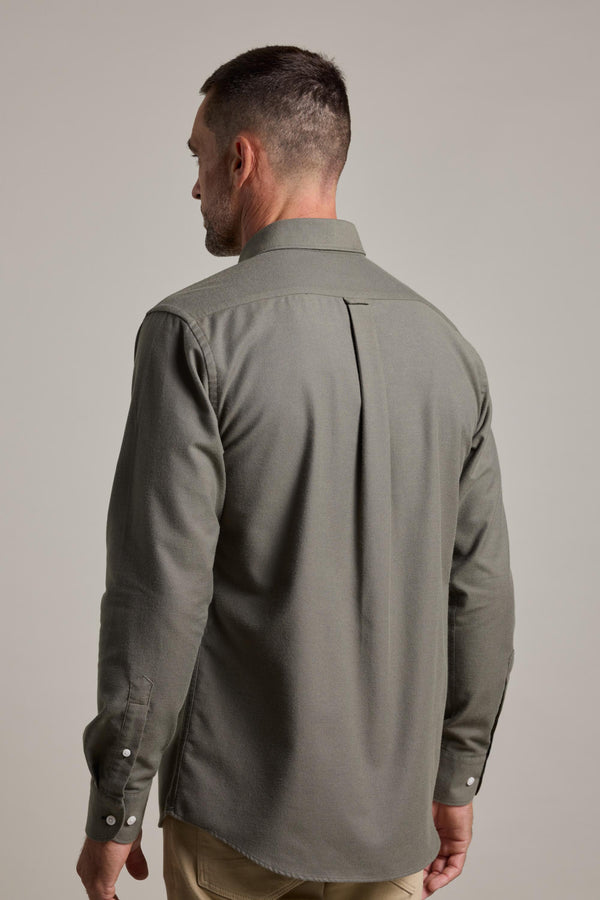 A man with short dark hair, seen from behind, wears a long-sleeved olive green Barkers Maynard Oxford Shirt and beige pants, standing against a plain light gray background.