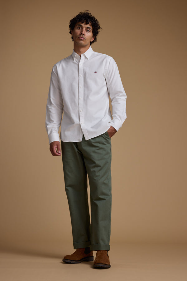 A man with curly hair stands against a tan background in a classic fit white Maynard Oxford Shirt by Barkers, paired with sage green trousers and brown suede shoes. He looks slightly upward in a relaxed pose.