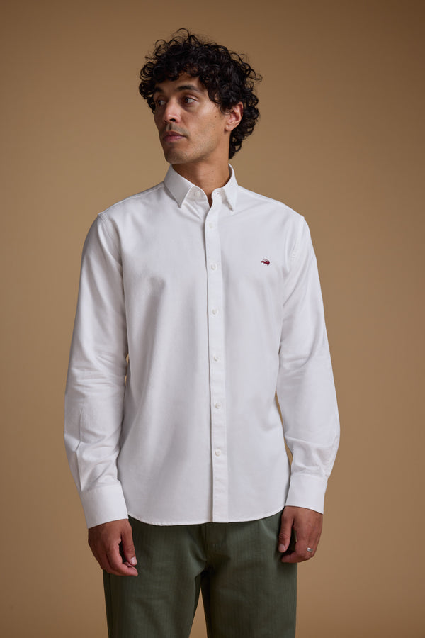 A man with curly hair wears a Barkers Maynard Oxford Shirt and olive green pants, standing against a plain beige background and gazing slightly left with a neutral expression.