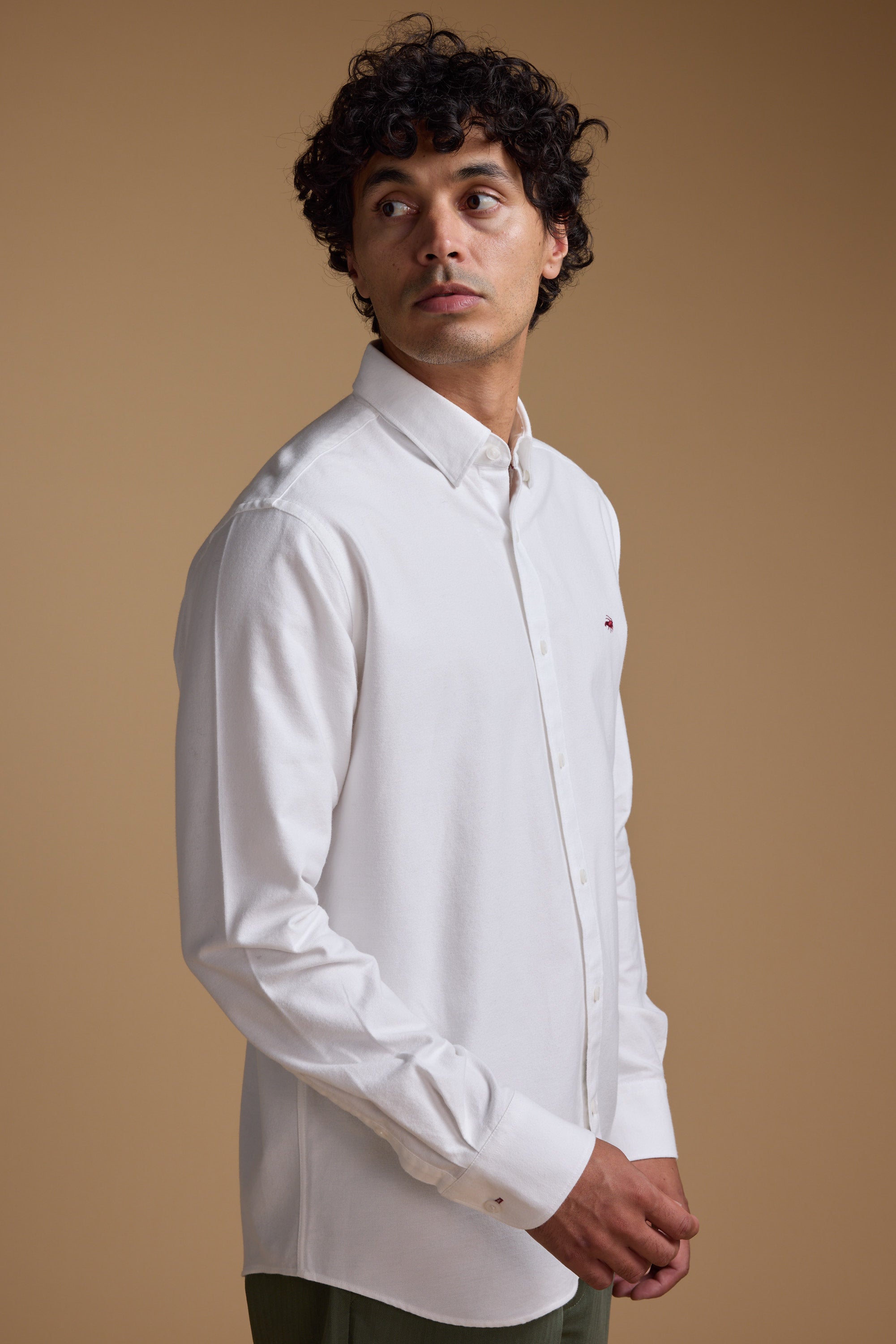 A man with curly dark hair wears a classic fit Barkers Maynard Oxford Shirt in white, made from responsibly sourced cotton, standing against a plain beige background and looking slightly upward to his left with a neutral expression.