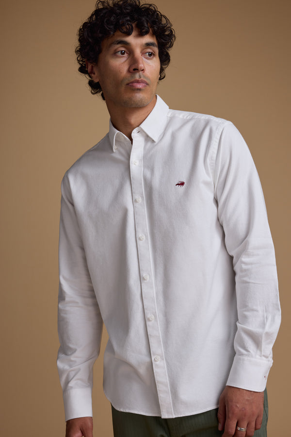 A man with curly hair wears a white Barkers Maynard Oxford Shirt, crafted from responsibly sourced cotton, standing against a plain beige background.