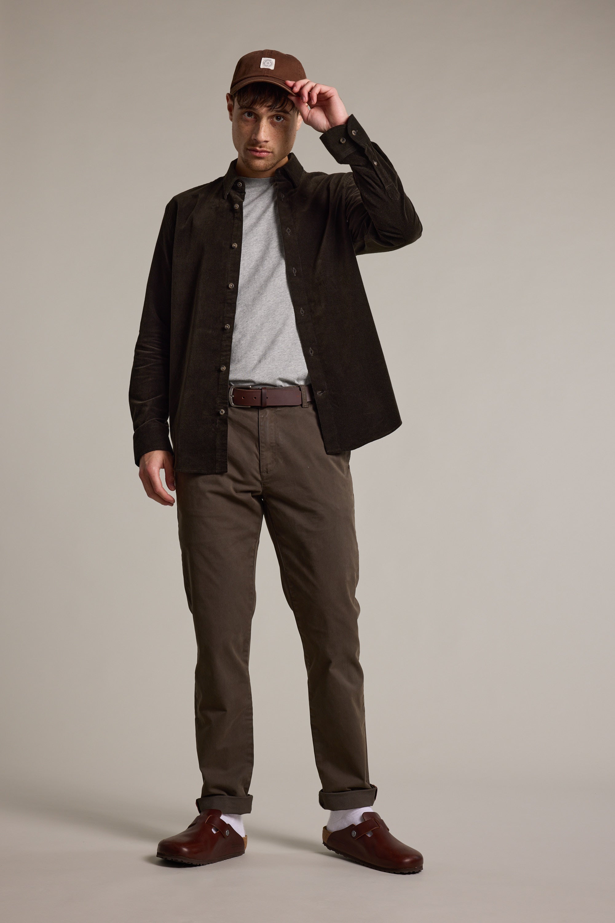 A man stands against a plain background, wearing the Barkers Washed Cotton Cap by Barkers, a dark brown button-up shirt over a gray t-shirt, brown belted pants, white socks, and brown shoes. He holds the cap’s brim with one hand.