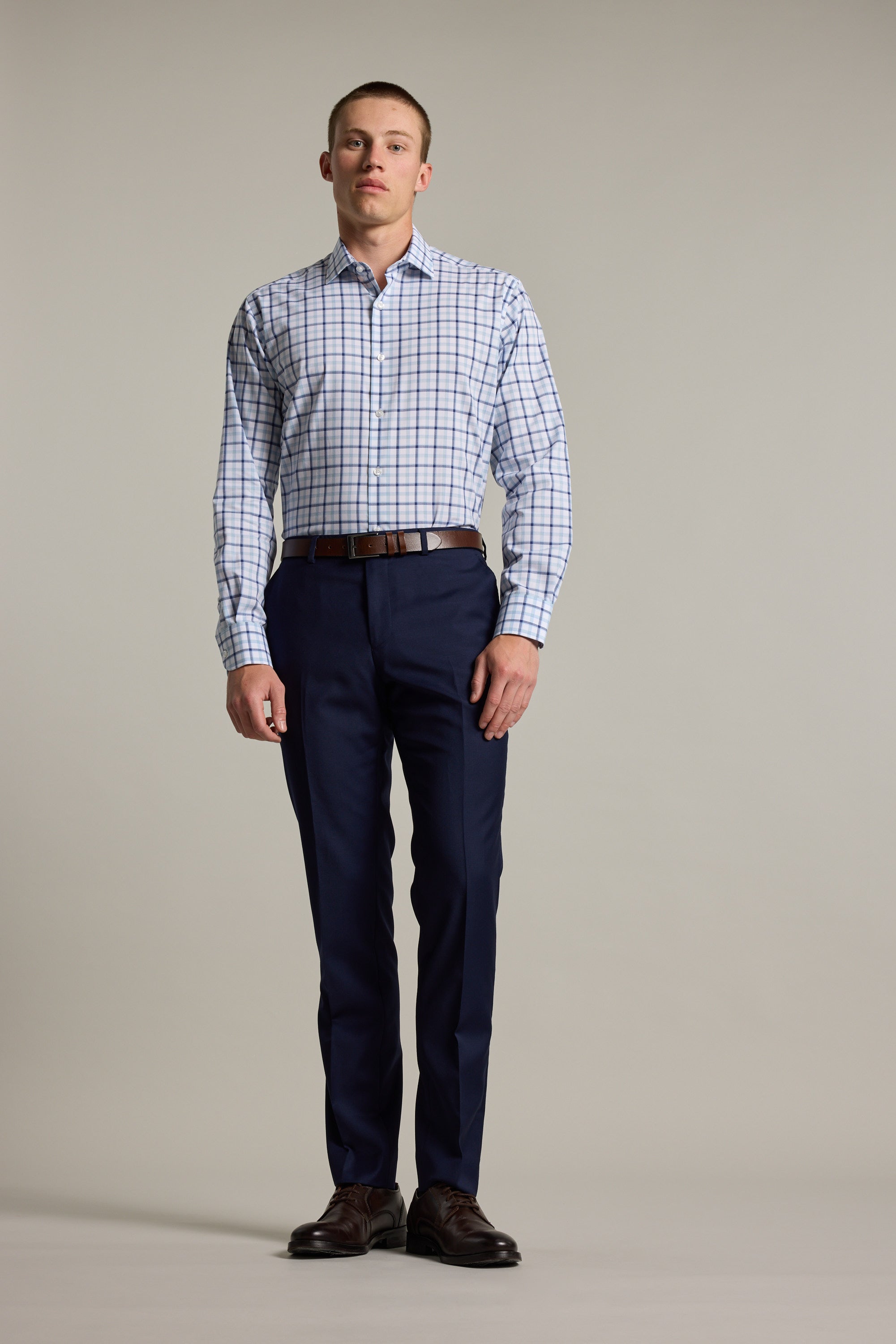 A man stands against a plain background wearing the Barkers Wesley Check Business Shirt paired with dark blue dress pants, a brown belt, and dark brown dress shoes.