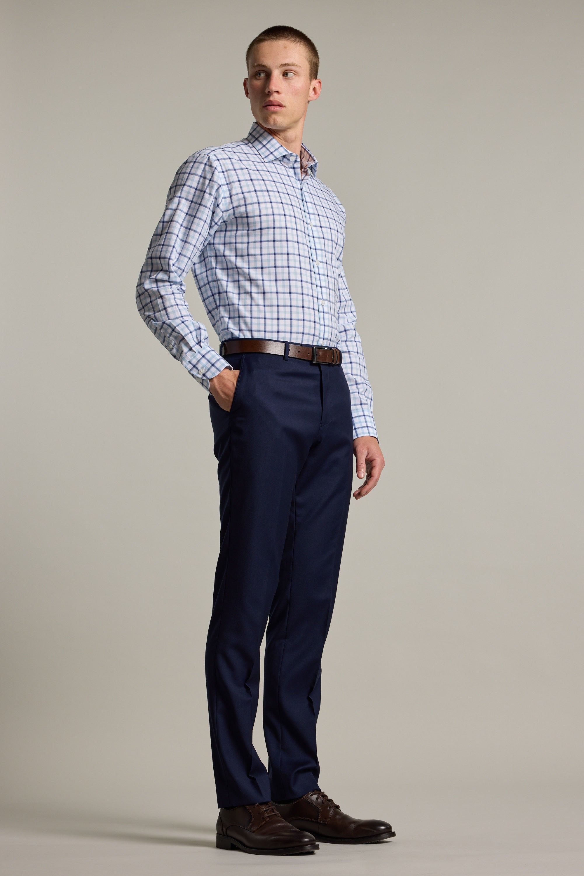 A man stands against a plain background wearing the Barkers Wesley Check Business Shirt. He pairs it with navy dress pants, a brown belt, and brown dress shoes, looking slightly to the side with a neutral expression.
