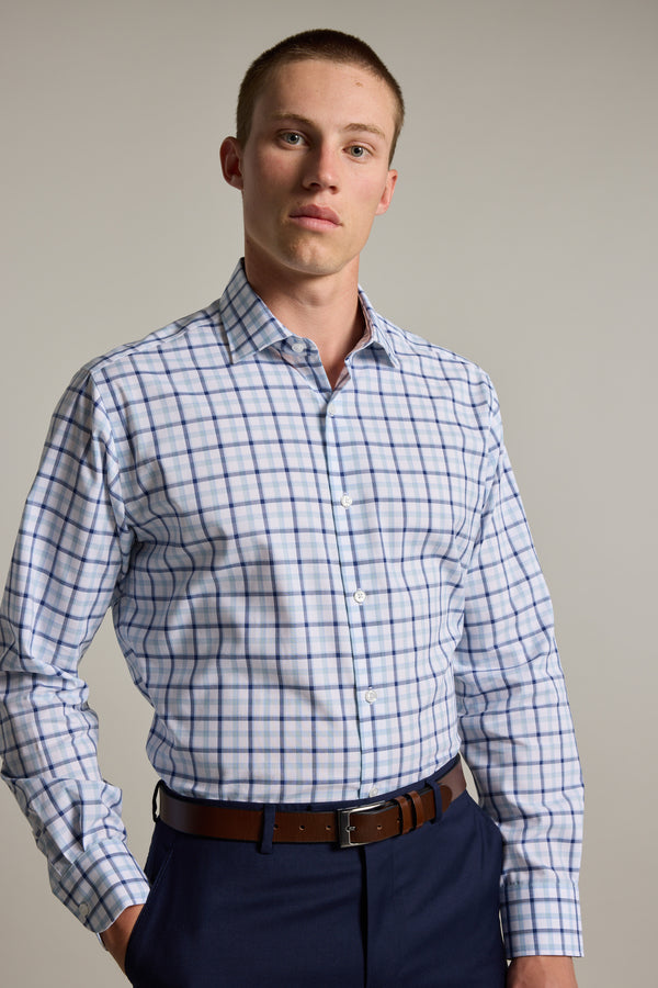 A young man with short hair wears the Barkers Wesley Check Business Shirt—a light-colored, long-sleeve, 100% cotton shirt with a blue and red plaid pattern—dark trousers, and a brown belt, standing against a plain background.