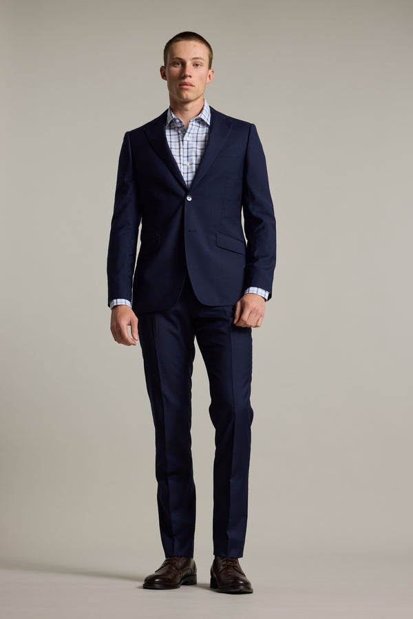 A man stands facing forward against a plain light gray background, wearing a navy blue suit paired with the Barkers Wesley Check Business Shirt in light blue and white, and dark brown dress shoes.