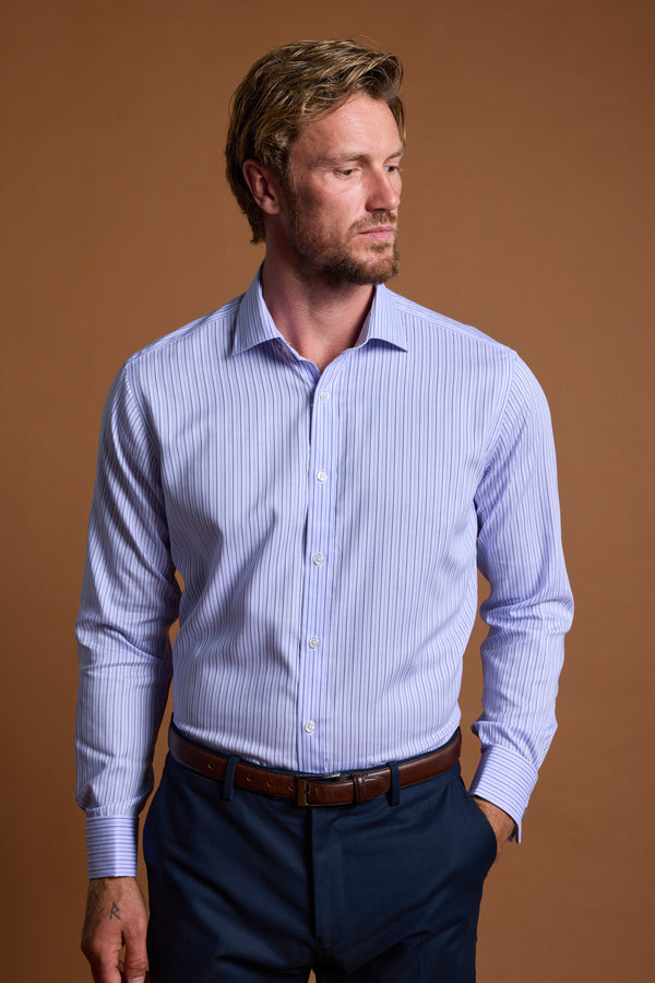 A man with short light brown hair and a beard wears the Barkers Asmara French Cuff Business Shirt, a tailored fit light blue vertically striped shirt, and dark blue pants. He stands against a plain brown background, looking left with a neutral expression.