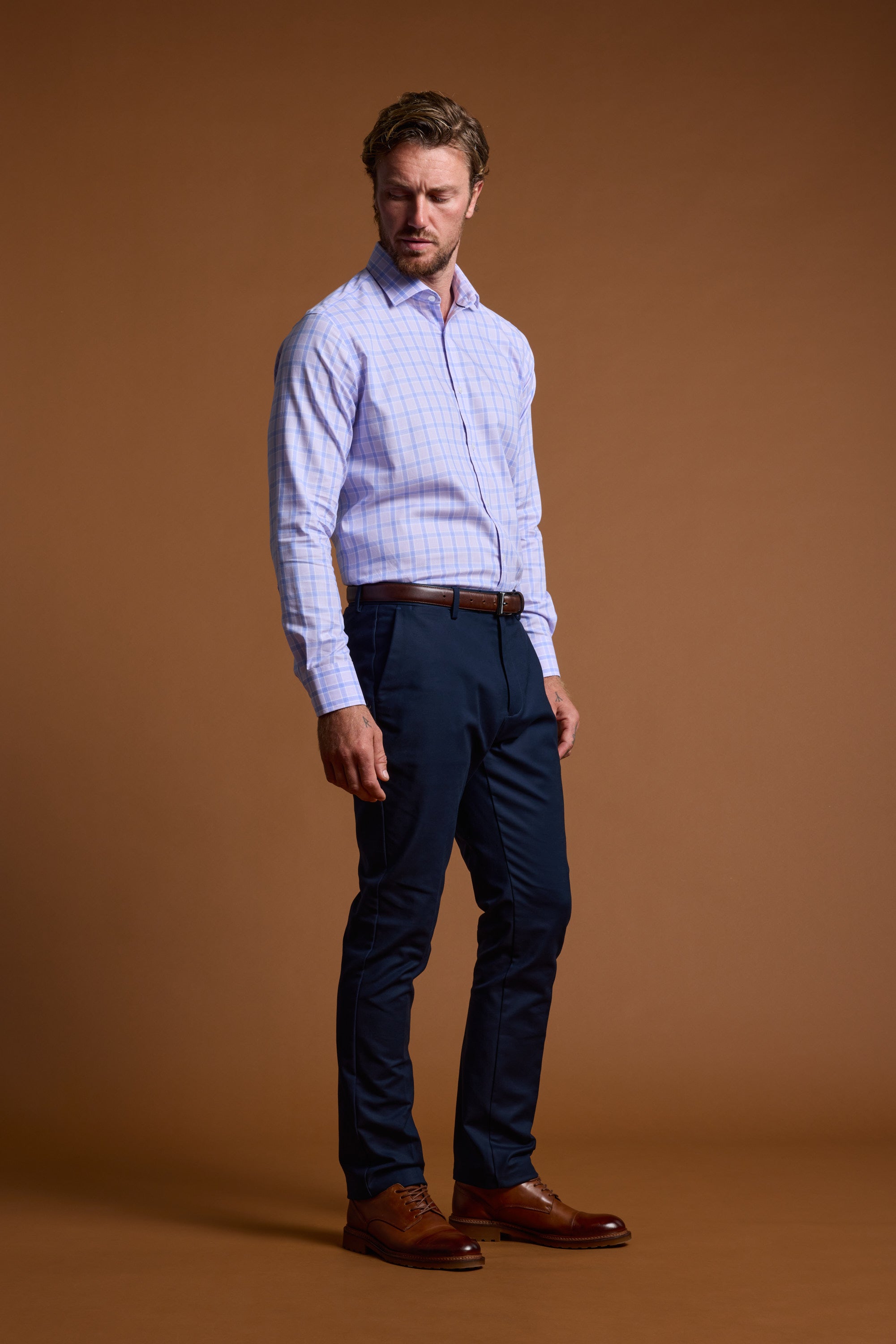 A man with short, wavy hair stands against a plain brown background wearing the Barkers Larry Check Business Shirt—light blue and wrinkle-free—paired with dark navy trousers, a brown belt, and matching dress shoes. He looks slightly to his left.