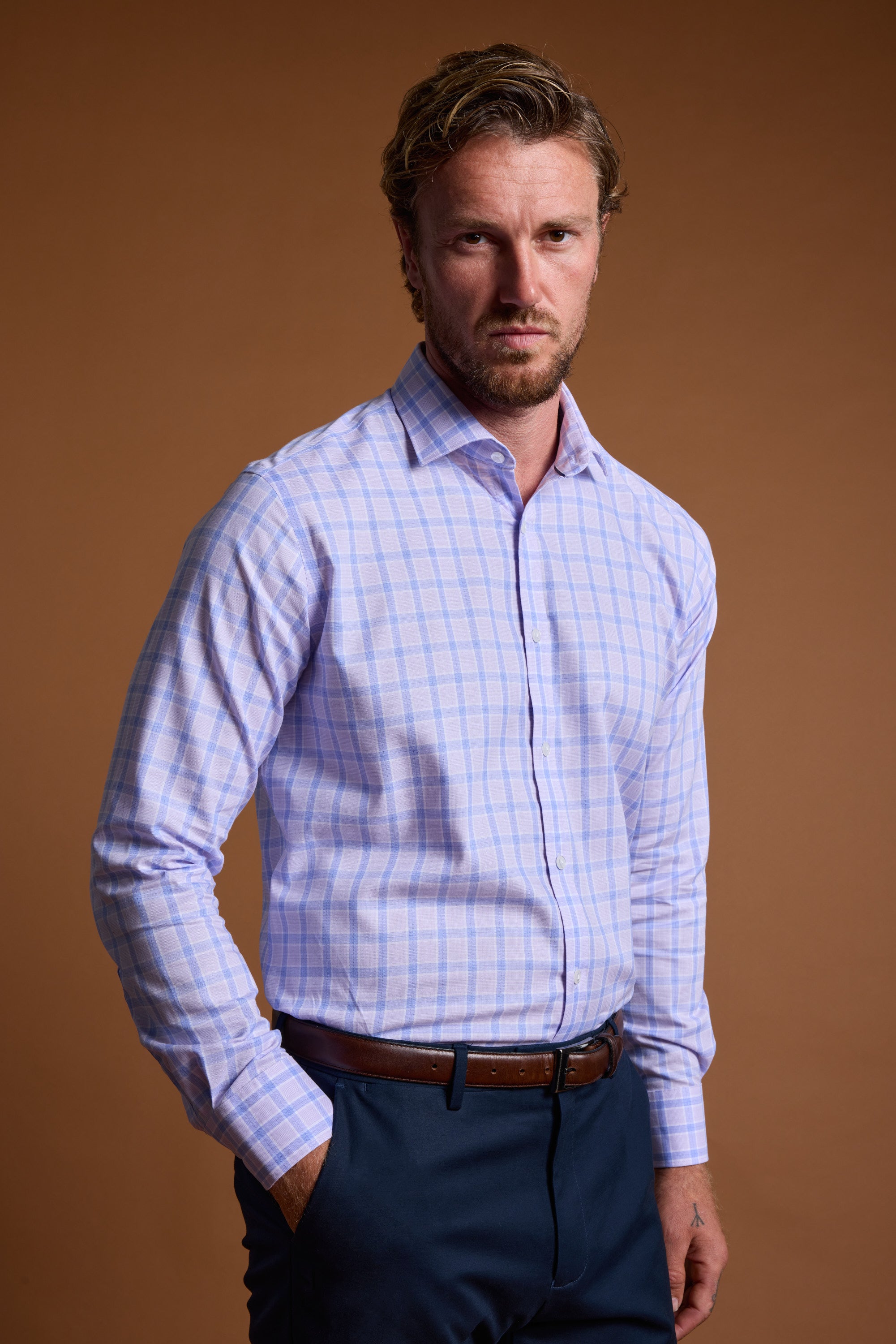A man with short, wavy hair and a beard wears the Barkers Larry Check Business Shirt tucked into navy pants with a brown belt, standing against a plain brown background with one hand in his pocket.
