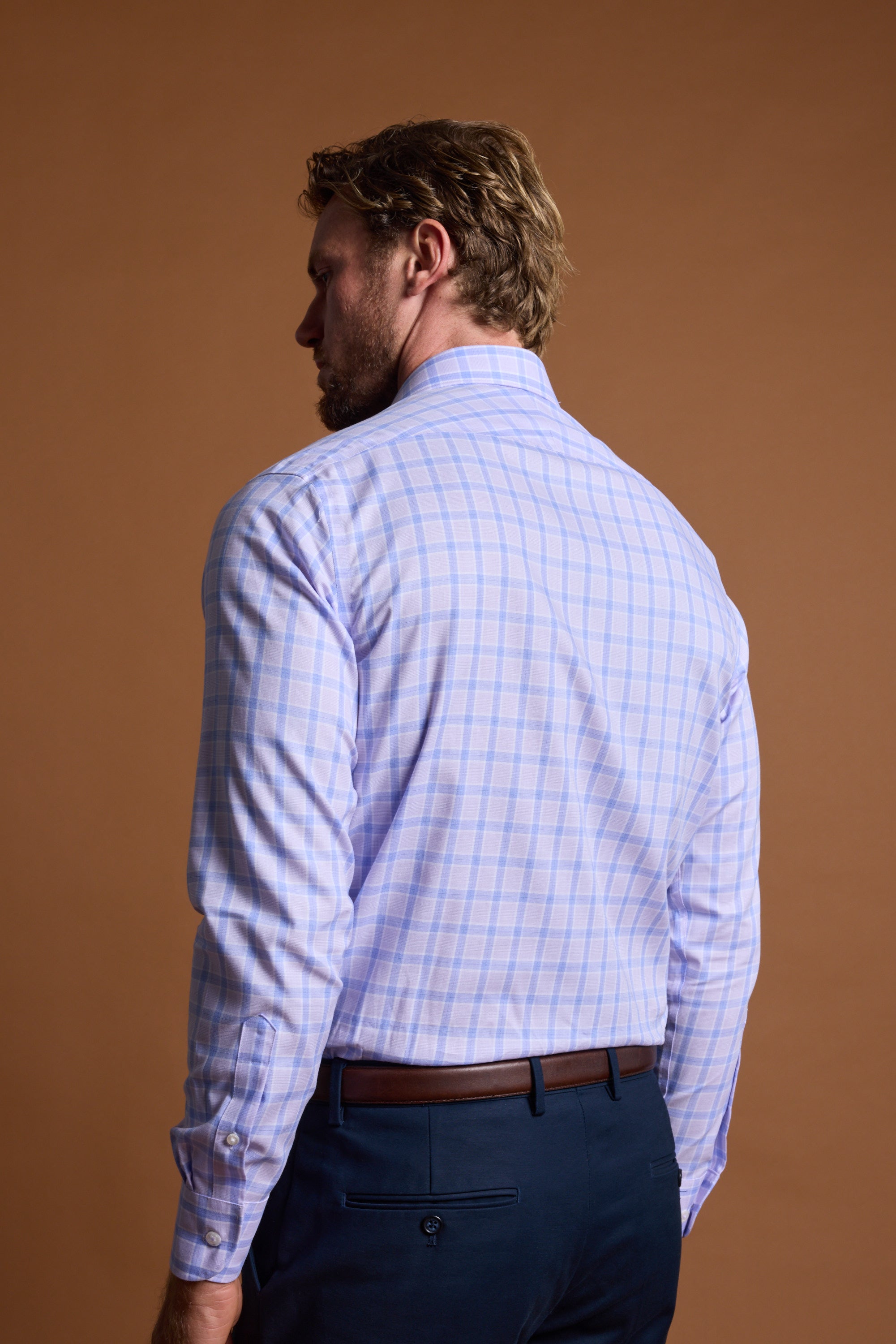 A man with light brown hair, seen from behind, wears the Barkers Larry Check Business Shirt—wrinkle-free plaid in light blue and pink—tucked into dark blue pants with a brown belt, standing against a plain brown background.
