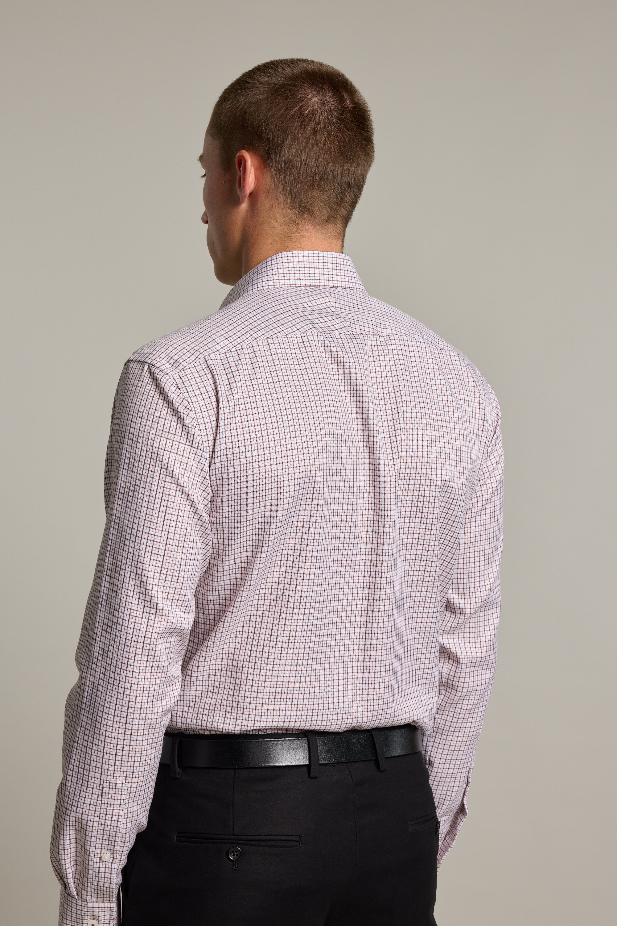 A man with short hair is seen from behind, wearing a light Marbury Check Business Shirt by Barkers and black dress pants with a belt, standing against a plain background.