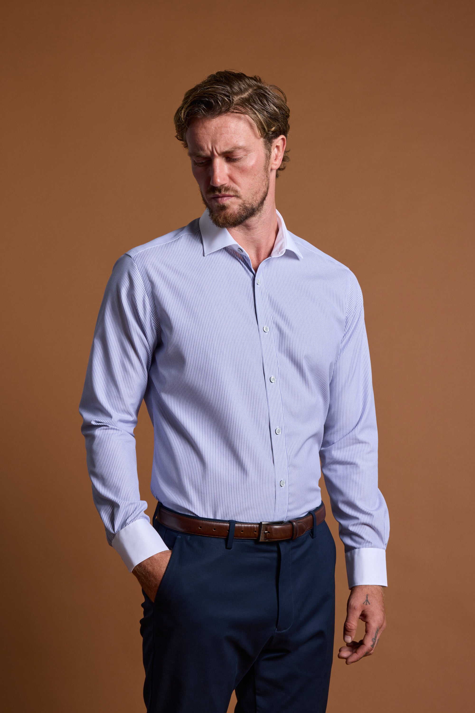 A man with light brown hair and a beard models the Barkers Felix Stripe Business Shirt in tailored fit with white cuffs, paired with navy trousers and a brown belt, showcasing Eco Easy Care style against a plain brown background.