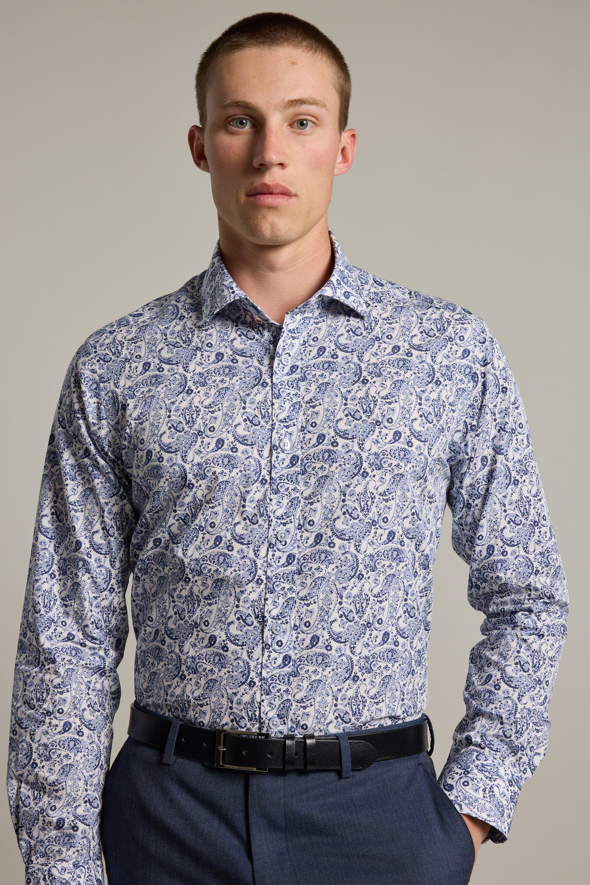 A young man with short hair wears the Buckman Print Business Shirt by Barkers, featuring a blue and white paisley pattern, paired with navy trousers and a black belt, standing against a plain light gray background.