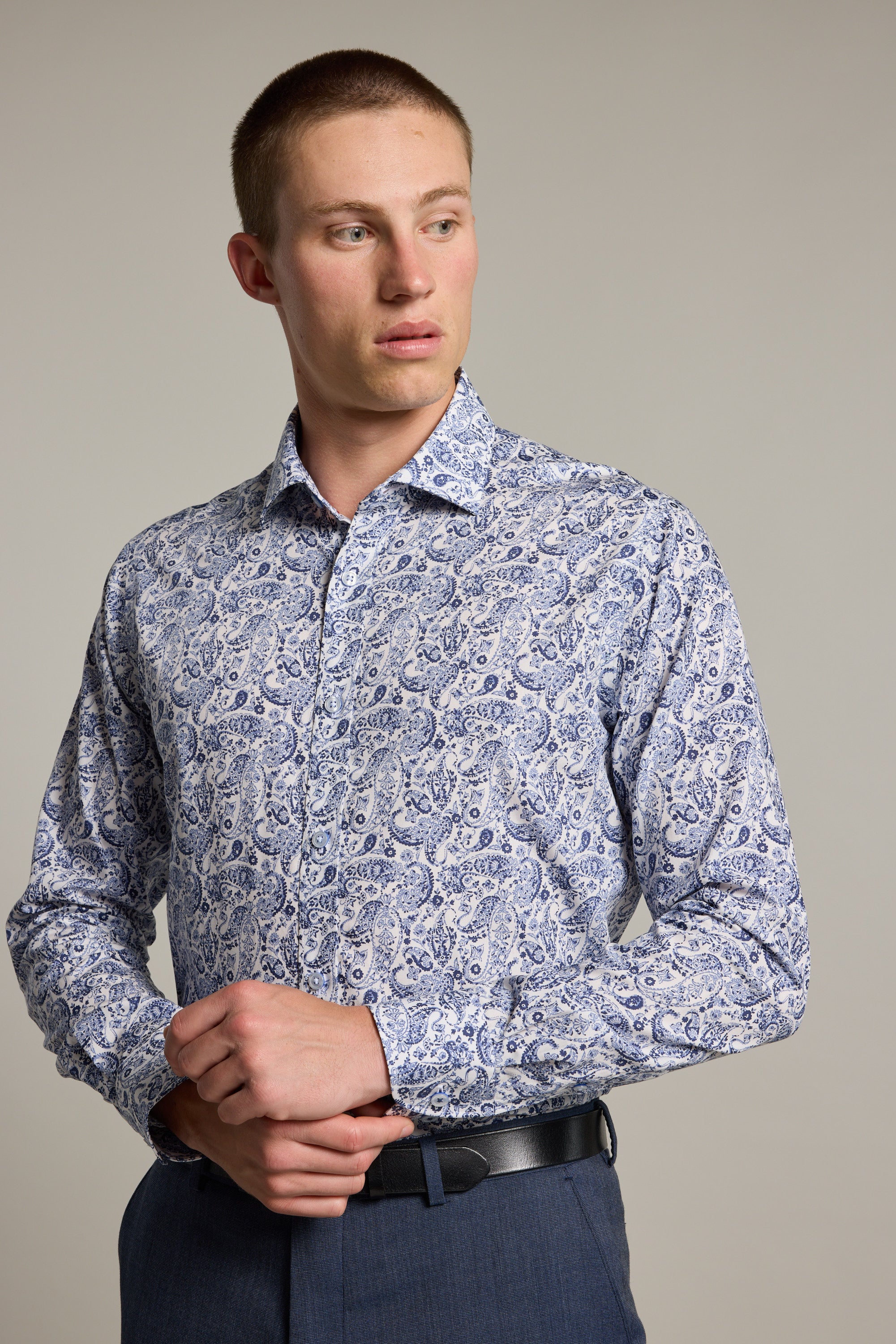 A man wears the Barkers Buckman Print Business Shirt, a tailored-fit, long-sleeve, light blue and white paisley-patterned shirt with dark pants. He looks left with his hands clasped near his waist against a neutral background.