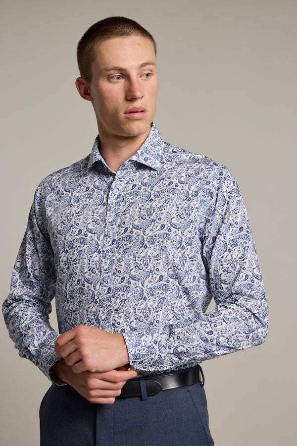 A man wears the Barkers Buckman Print Business Shirt, a tailored-fit, long-sleeve, light blue and white paisley-patterned shirt with dark pants. He looks left with his hands clasped near his waist against a neutral background.