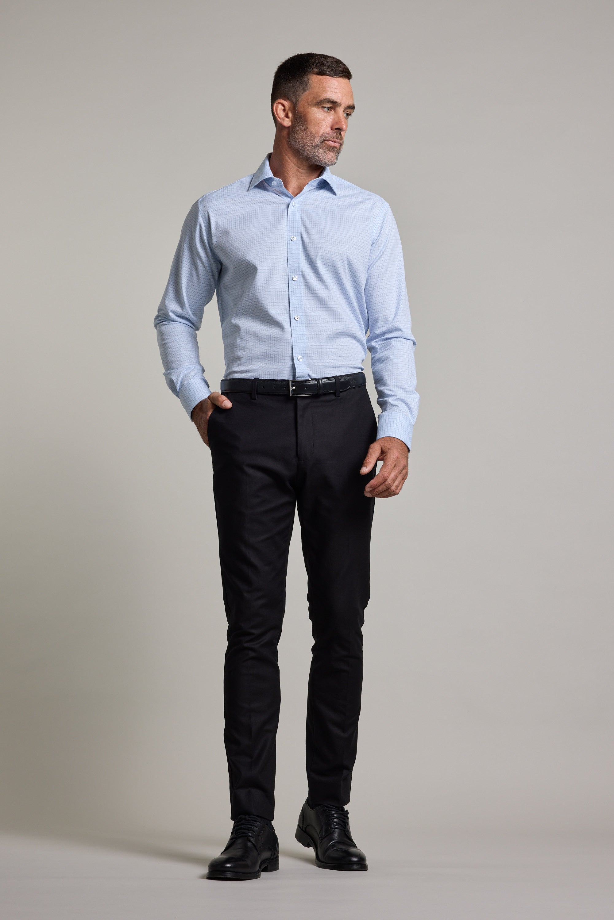 A man stands against a plain background, wearing the Barkers Hunter Check Business Shirt, dark slim-fit trousers, a black belt, and black dress shoes. He has one hand in his pocket and looks to the side.