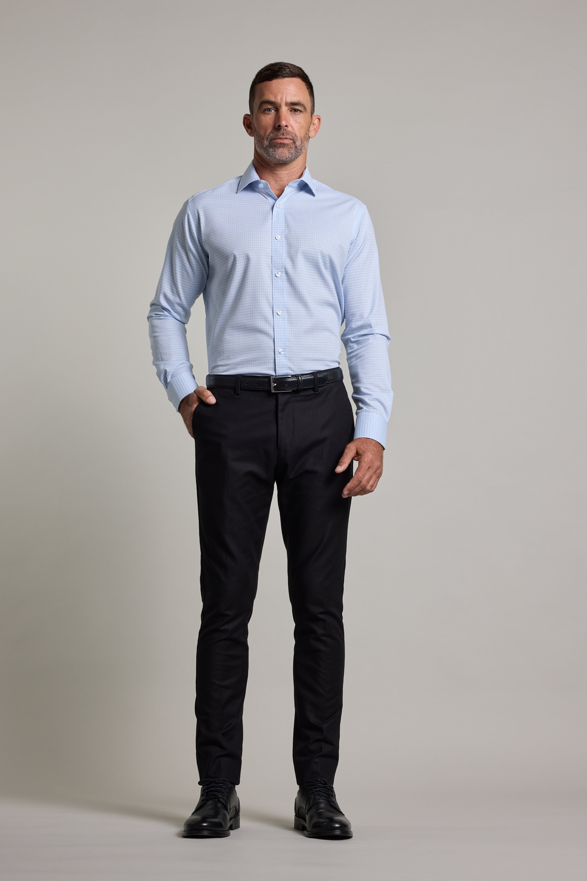 A man stands facing forward against a plain background, wearing the Barkers Hunter Check Business Shirt with black pants, a belt, and black dress shoes. One hand is in his pocket and he has a neutral expression.