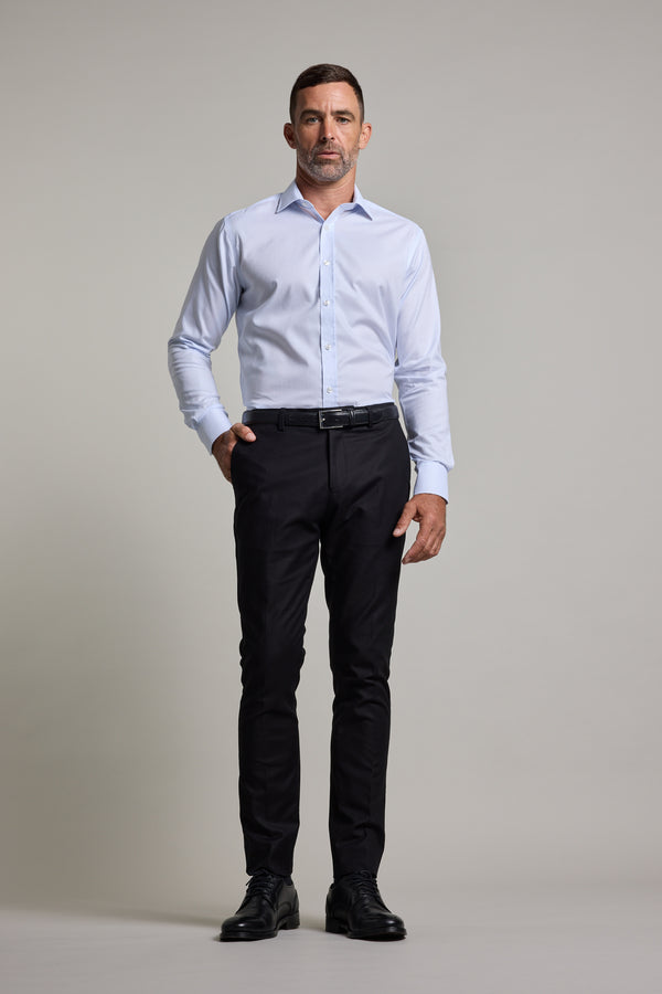 A man faces forward in a tailored fit Barkers Penny Texture Business Shirt, paired with dark dress pants and black shoes, one hand in his pocket against a plain light background.