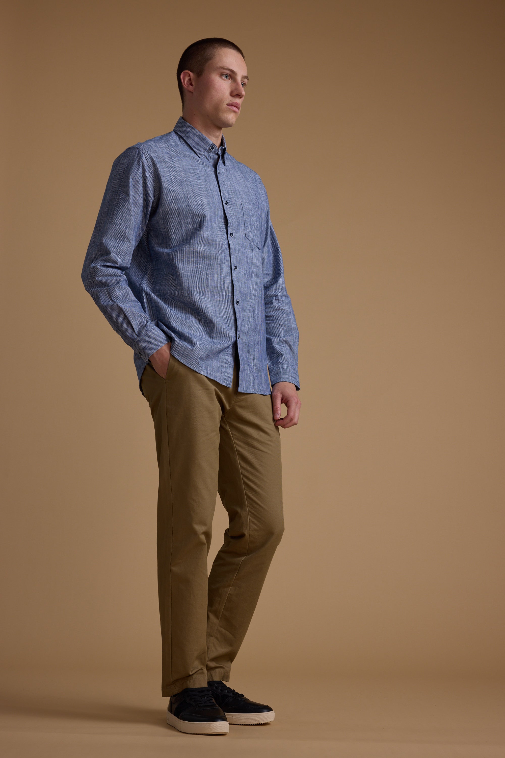 A man stands against a beige background wearing the Blaine Stripe Shirt by Barkers, made from responsibly sourced cotton, paired with tan pants and black shoes. He has his left hand in his pocket and looks slightly to the side.