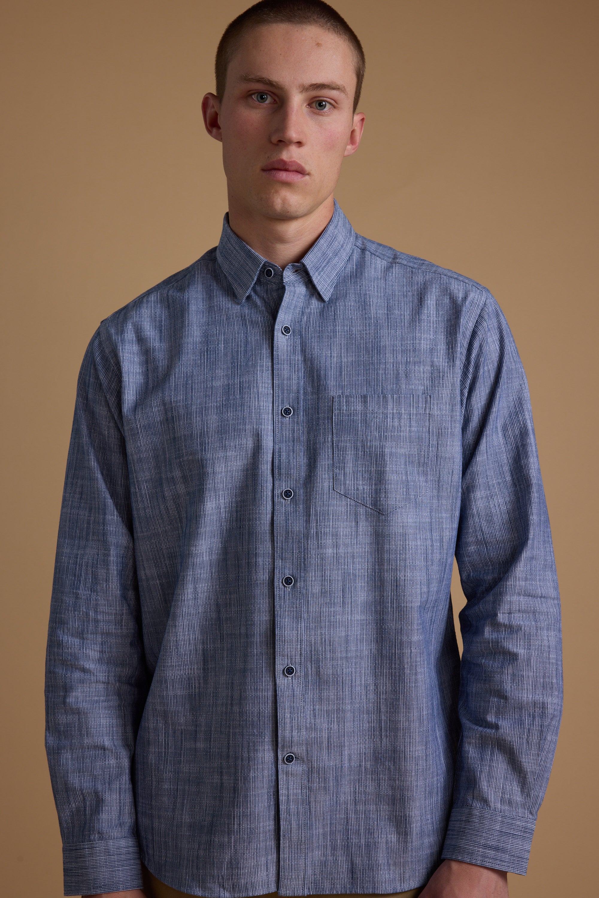 A young man with short hair wears the Barkers Blaine Stripe Shirt, a classic fit made from responsibly sourced cotton, featuring blue stripes. He stands against a plain beige background, looking directly at the camera with a neutral expression.