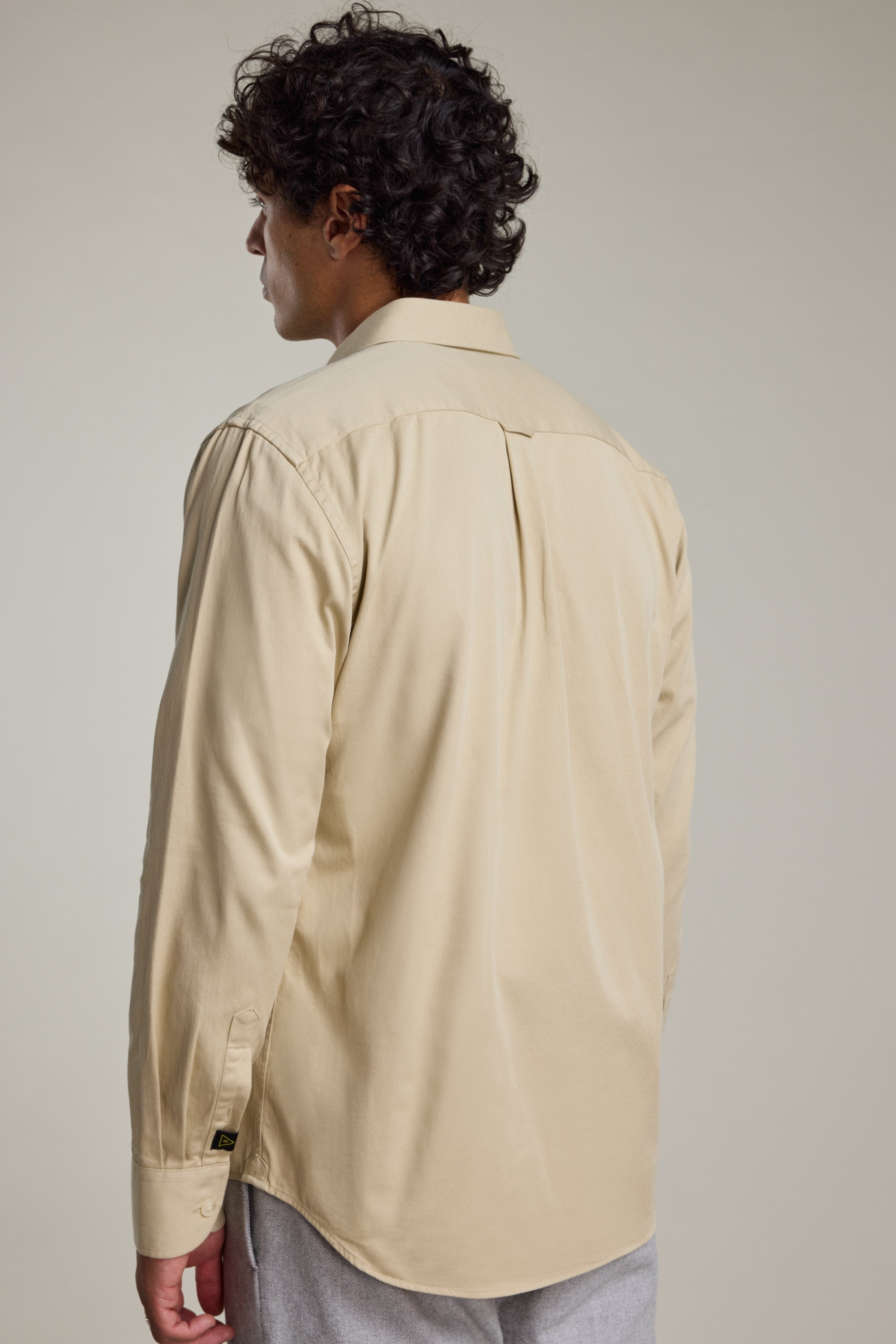 A man with curly dark hair is seen from behind, wearing the Barkers Chadwick Overshirt in beige and light gray pants against a neutral background—a true layering essential.