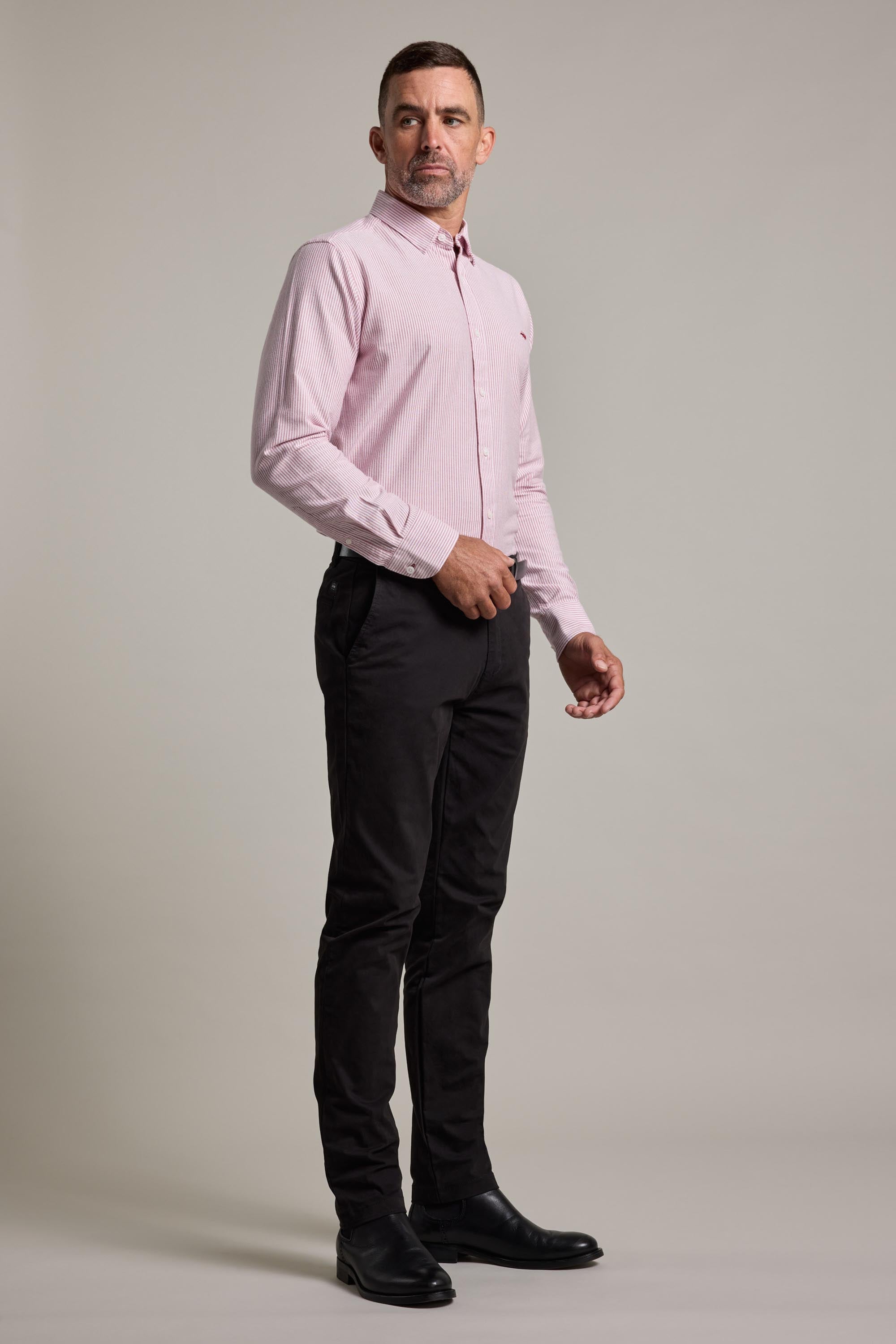 A man stands against a plain background, wearing the Barkers Maynard Oxford Stripe Shirt, black pants, and black shoes. He faces forward with a neutral expression and his right hand near his waist.