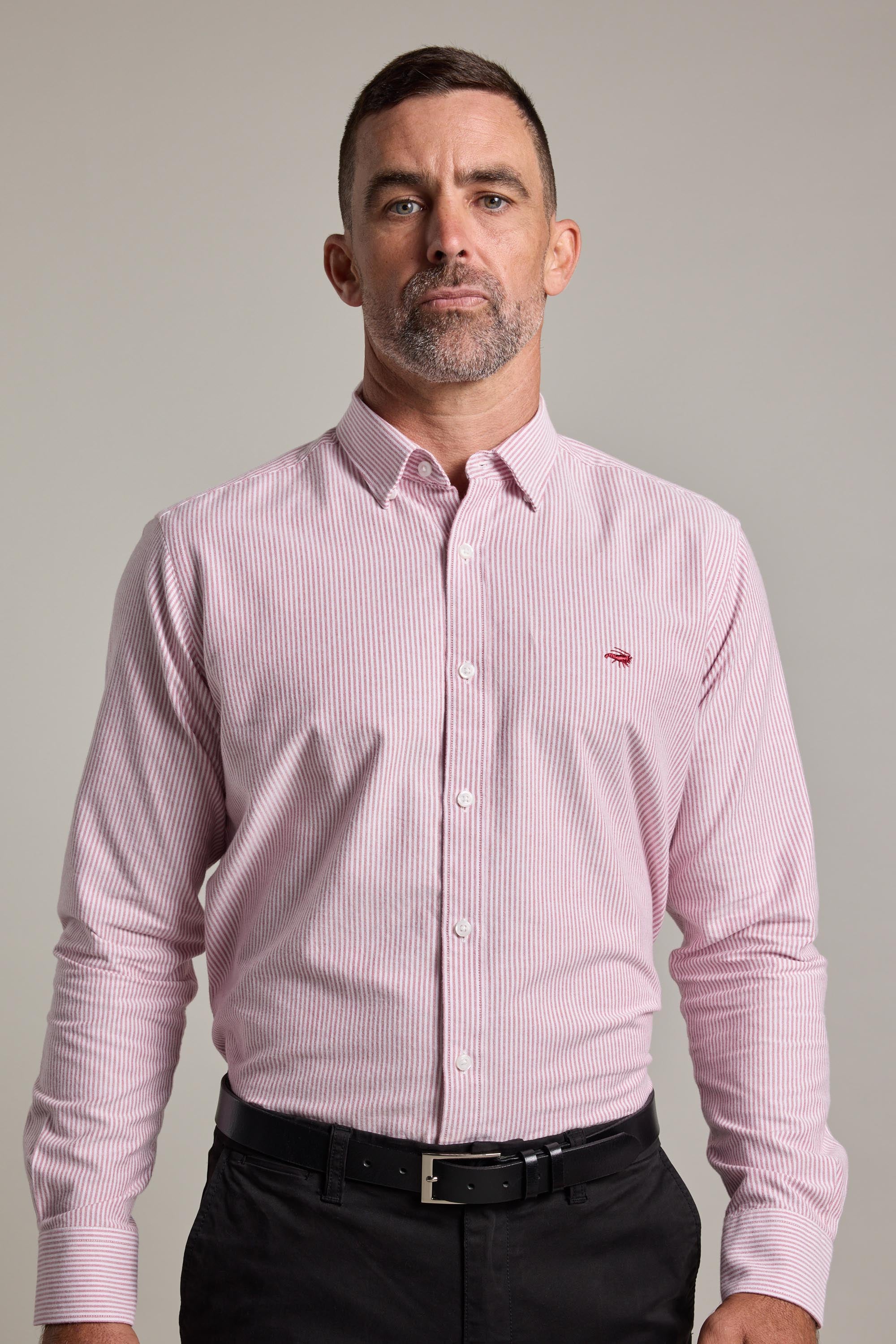 A man with short hair and a trimmed beard wears the Barkers Maynard Oxford Stripe Shirt, a light pink, classic fit men's cotton shirt with thin stripes, tucked into black pants and paired with a black belt against a neutral background.