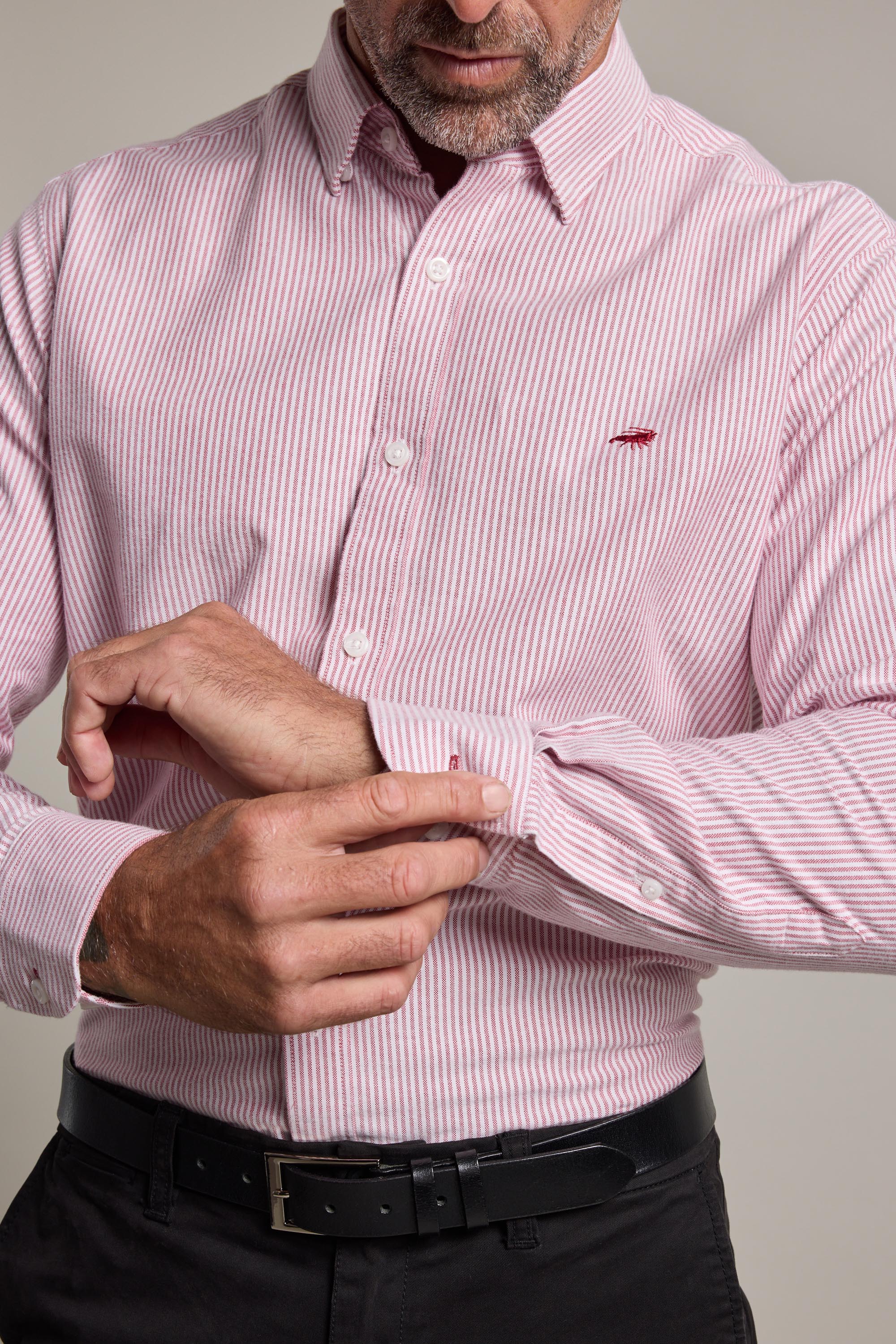 A man adjusts his left cuff while wearing the Barkers Maynard Oxford Stripe Shirt in pink and white, featuring a classic fit, small red chest logo, and tucked into black pants with a black belt.