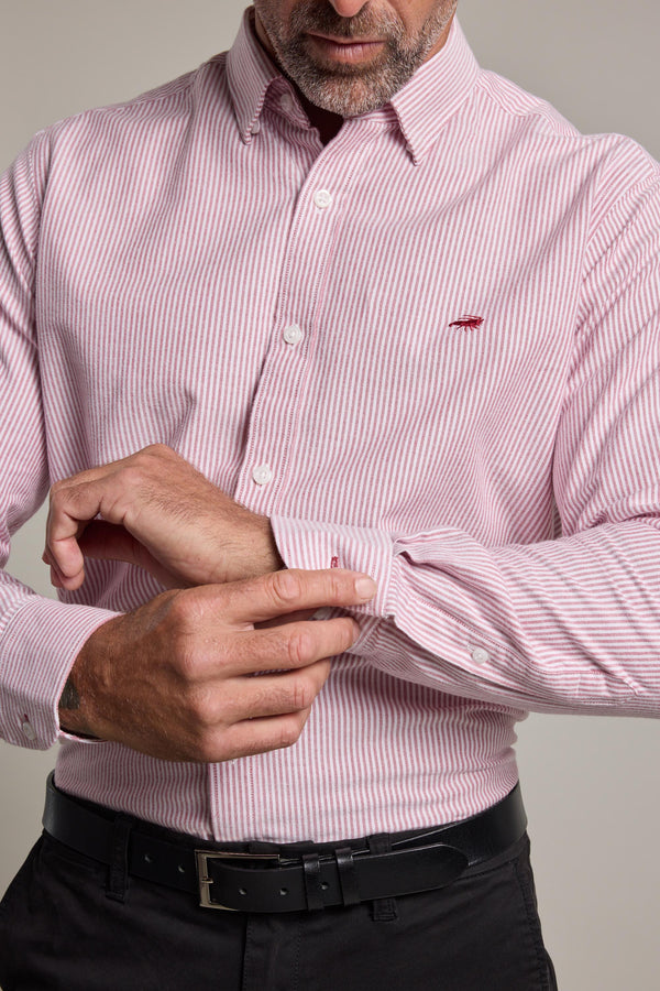 A man adjusts his left cuff while wearing the Barkers Maynard Oxford Stripe Shirt in pink and white, featuring a classic fit, small red chest logo, and tucked into black pants with a black belt.