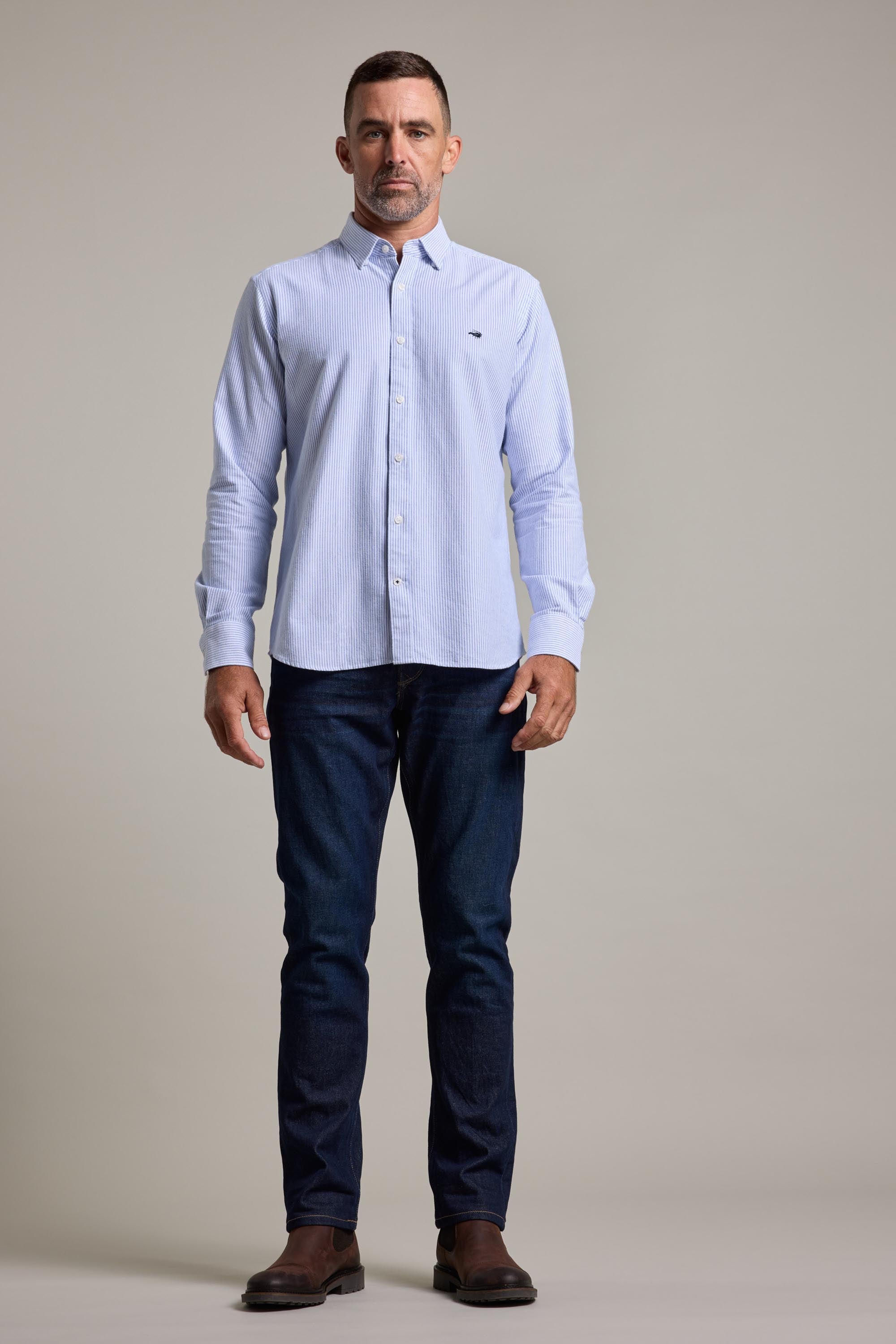 A man stands against a plain background wearing the Barkers Maynard Oxford Stripe Shirt, made from responsibly sourced cotton, paired with dark blue jeans and brown shoes. He has short hair, a neutral expression, and relaxed arms by his sides.