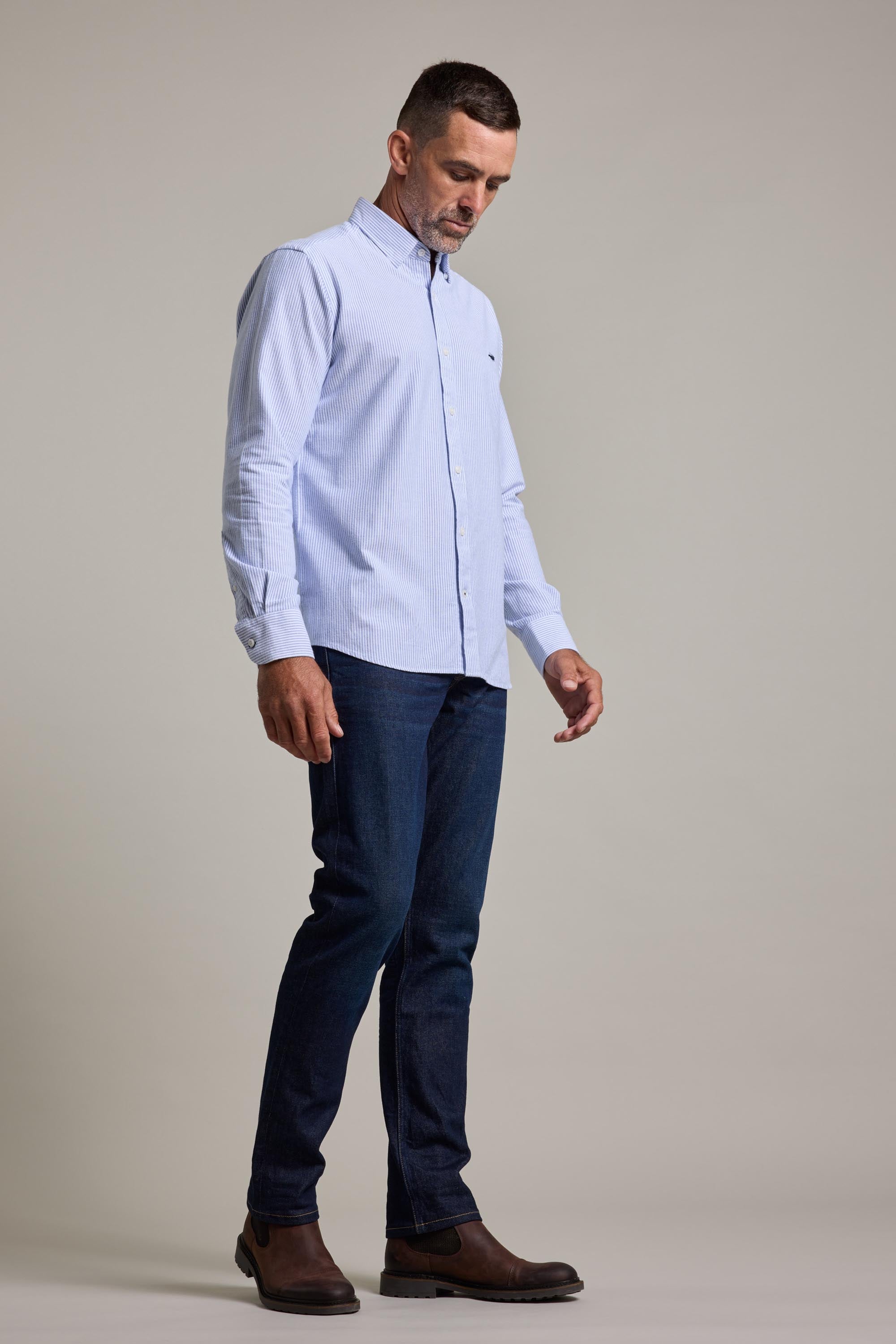 A man stands against a plain background, wearing the Barkers Maynard Oxford Stripe Shirt, dark blue jeans, and brown boots. He looks down slightly with his arms relaxed at his sides.