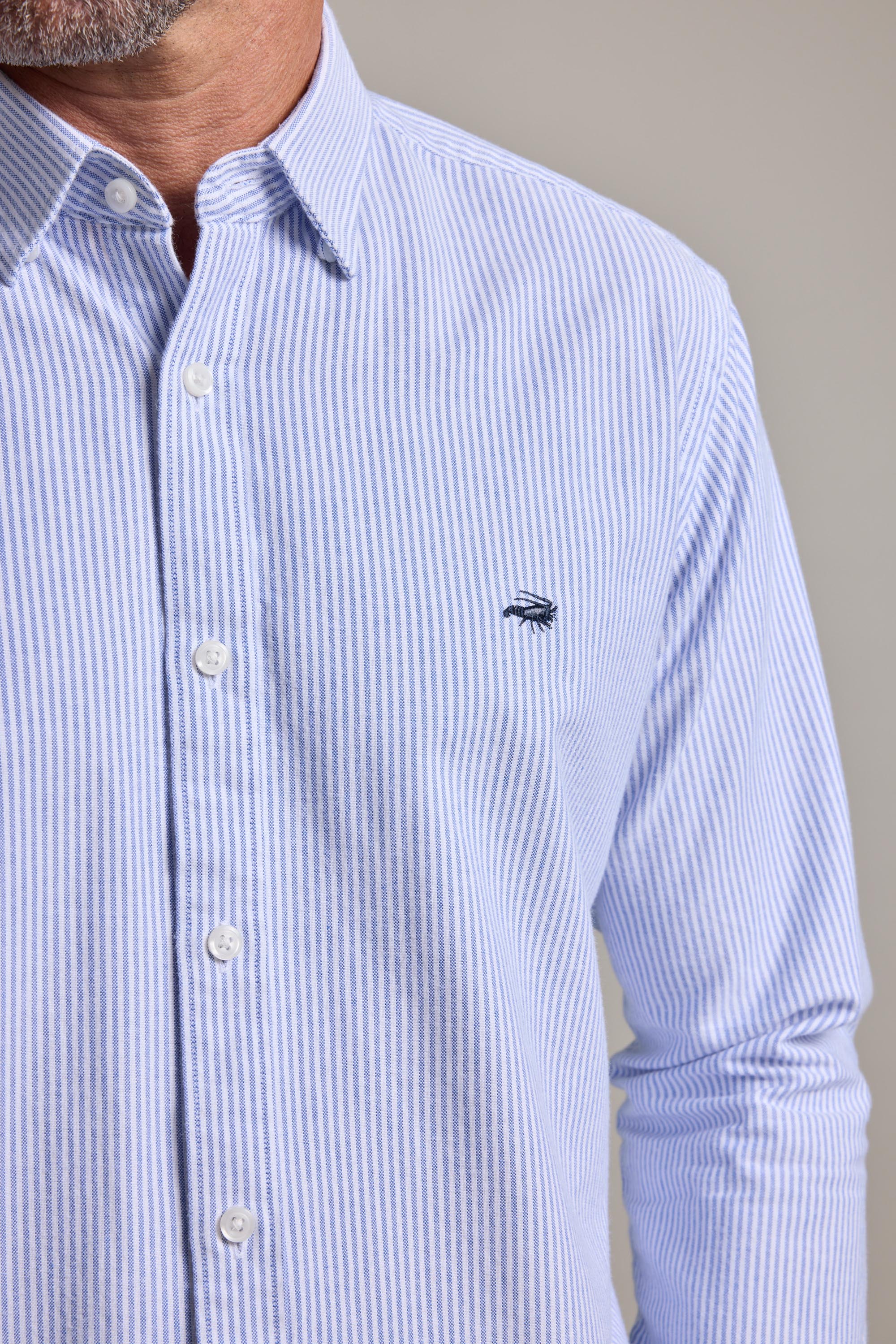 A man wears the Barkers Maynard Oxford Stripe Shirt, featuring light blue and white vertical stripes, responsibly sourced cotton, and a small chest logo. The image shows the men's classic shirt from shoulders to waist.