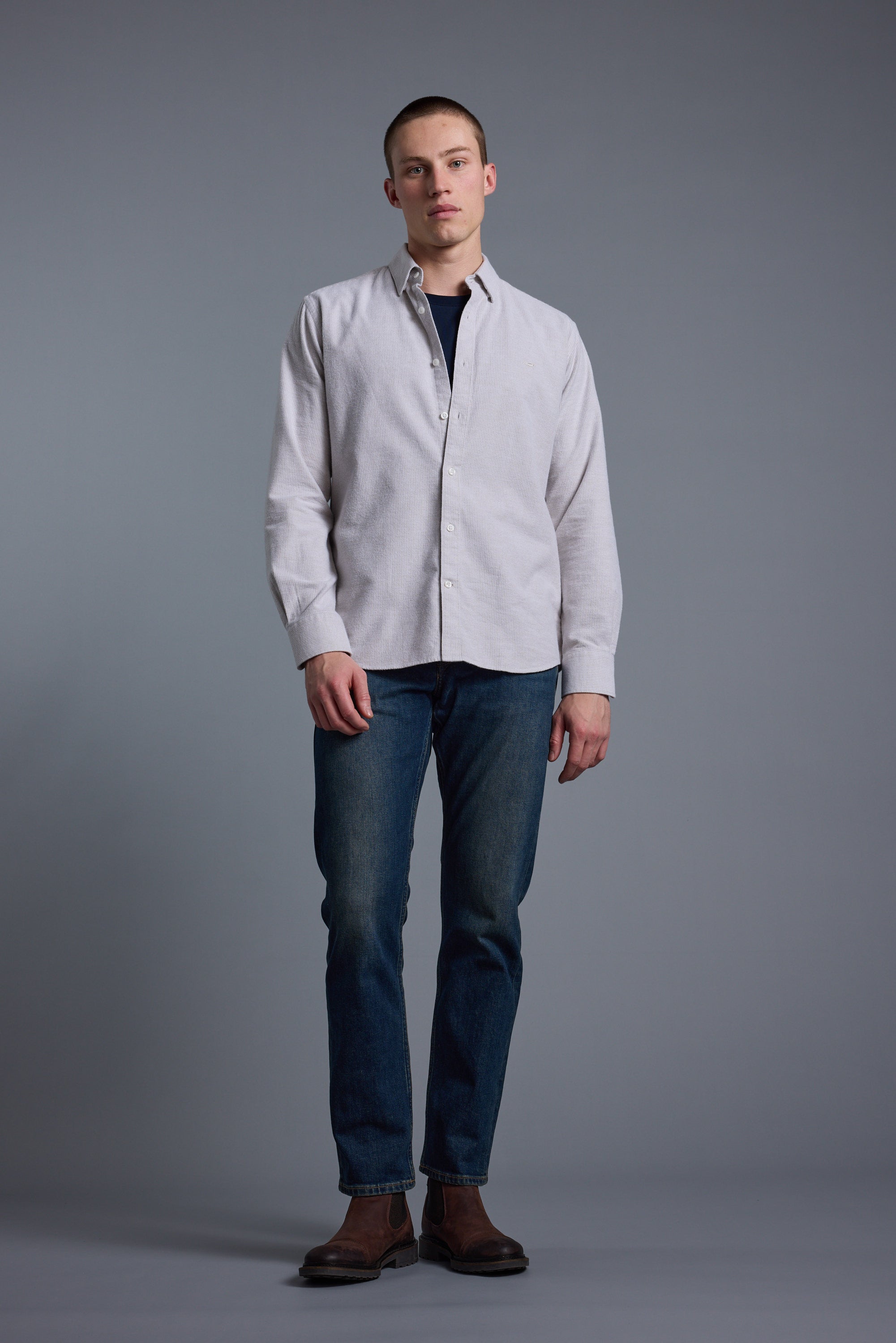 A person stands against a plain gray background wearing the Barkers Maynard Oxford Stripe Shirt over a dark shirt, paired with blue jeans and brown boots, looking directly at the camera with a neutral expression.