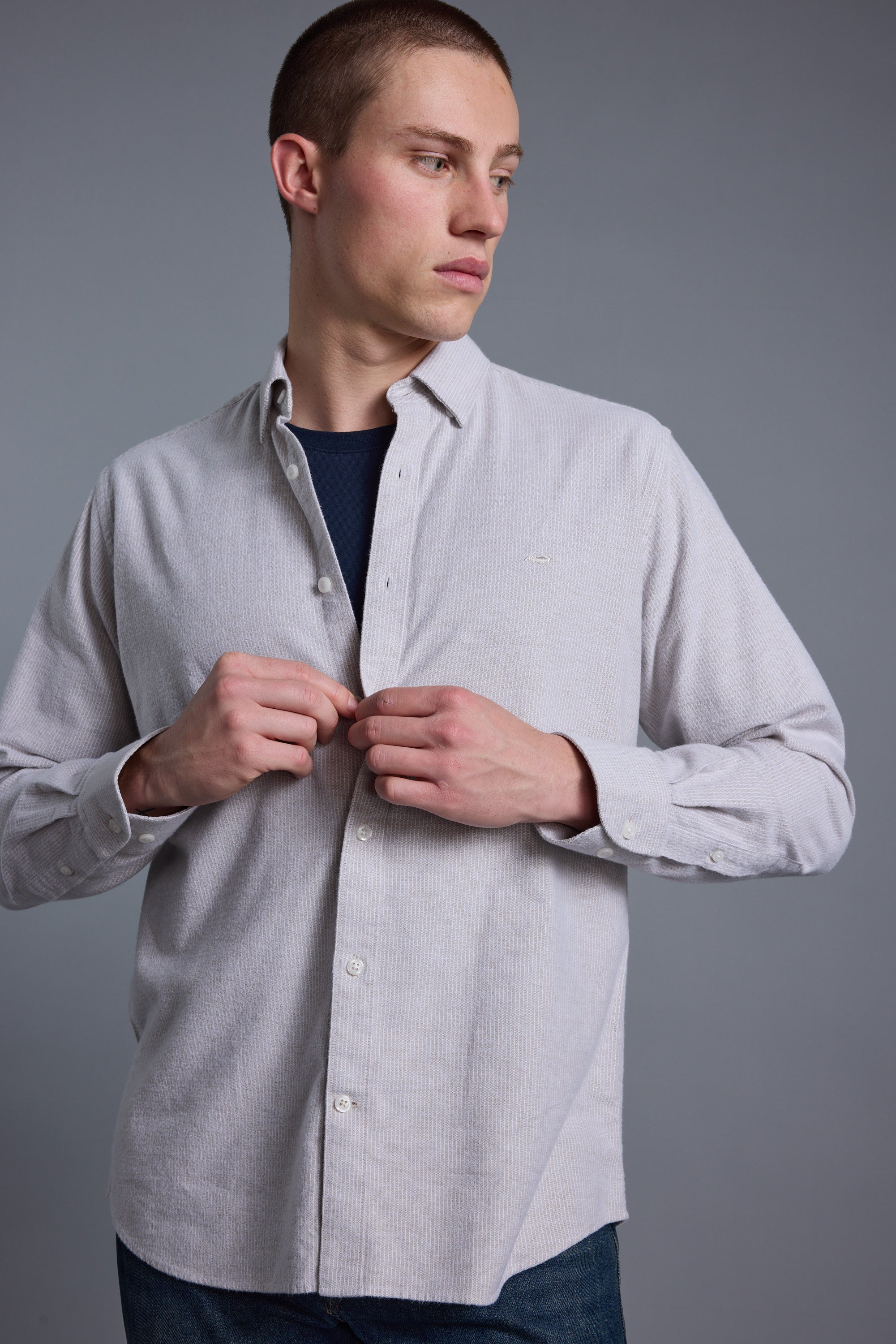A young man with short hair wears the Barkers Maynard Oxford Stripe Shirt, made from responsibly sourced cotton, over a dark tee. He stands against a plain gray background, looking left and fastening a button on his shirt.